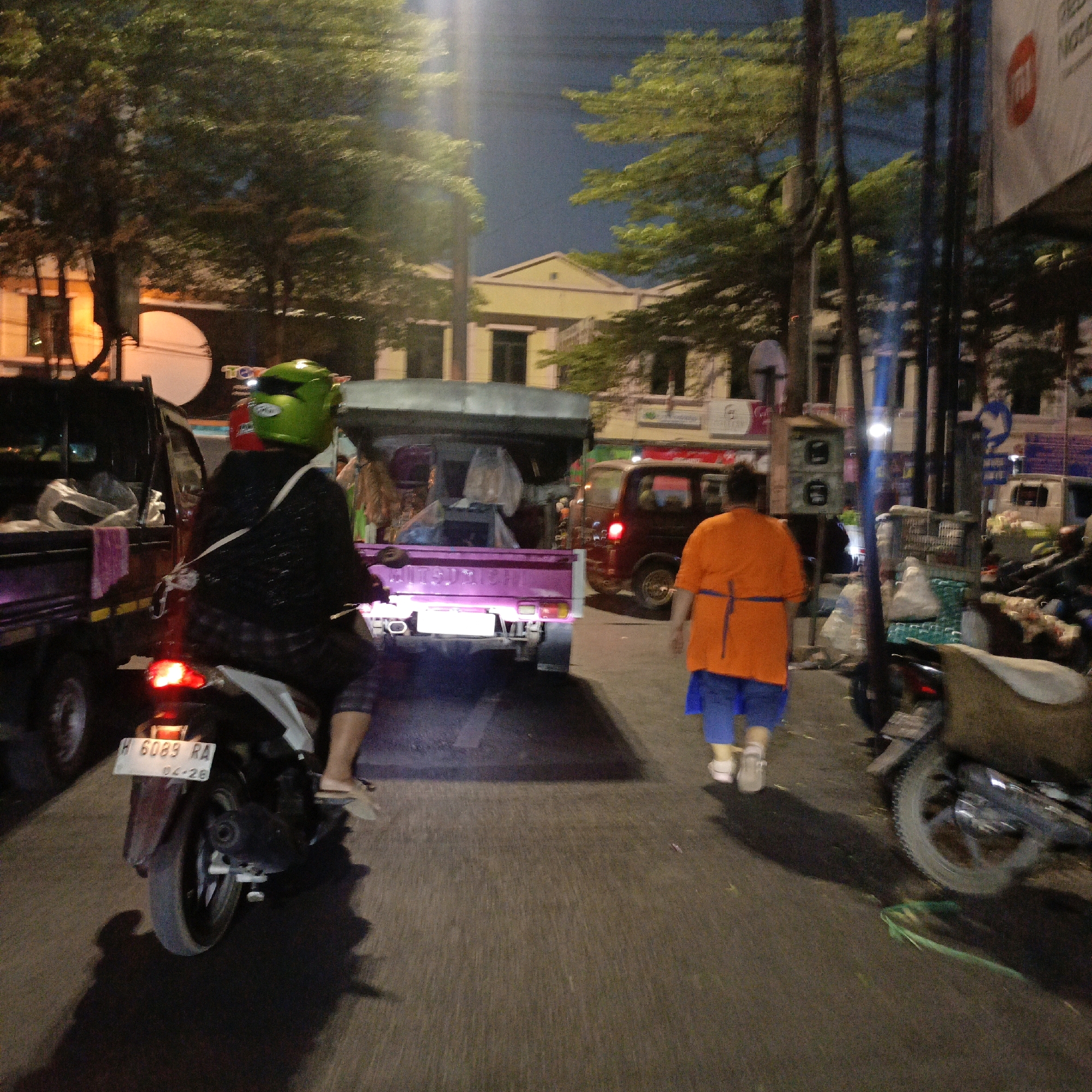 The image depicts a street scene at night in Salatiga, Indonesia. In the foreground, a person wearing a green helmet and a black jacket is riding a motorcycle with a license plate reading "H 6089 RA" and "04-26". The motorcycle has a red taillight illuminated. To the left, a dark-colored truck bed is visible, partially loaded with what appears to be plastic sheeting. In the middle ground, a purple pickup truck is parked, its rear taillights visible. To the right, a person in an orange shirt and blue shorts walks away from the camera. Further in the background, a dark red van is parked. Buildings line the street, illuminated by artificial lights, and trees with dark foliage are present. The street surface is asphalt, and the overall lighting suggests nighttime conditions.