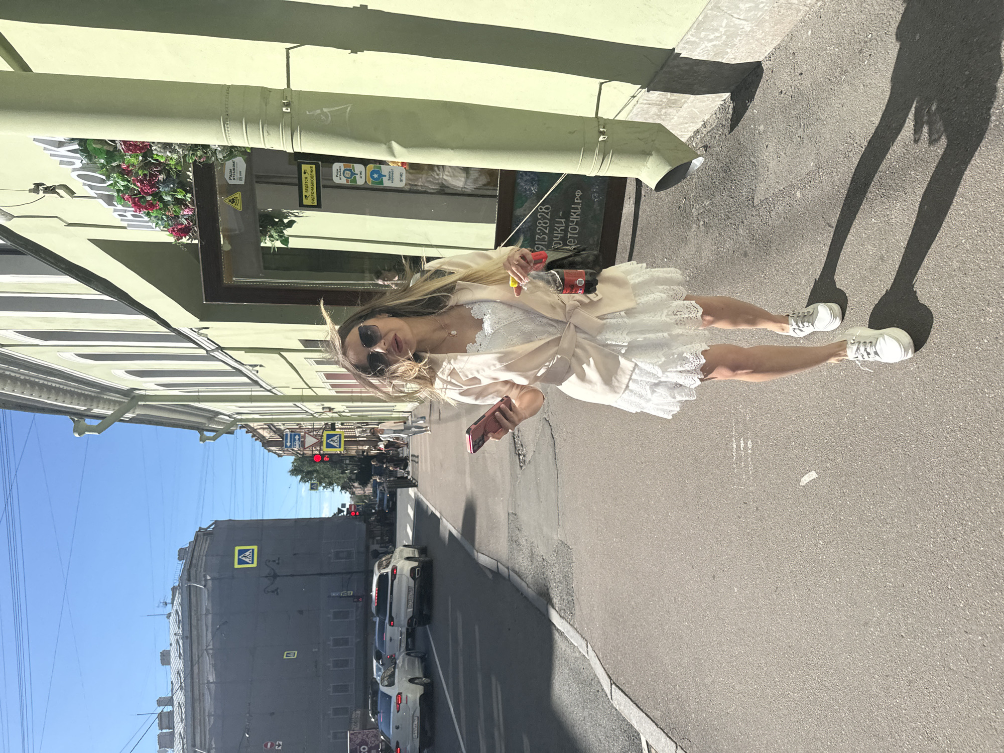 A young Caucasian woman with blonde hair and sunglasses walks down a sunlit sidewalk in Saint Petersburg, Russia, on a clear day. She is wearing a white lace dress with a light beige jacket and white sneakers. In her right hand, she holds a pink smartphone and a bottle of Coca-Cola. A dark backpack is slung over her left shoulder. The woman is walking towards the camera, on a sidewalk adjacent to a street with light traffic, including white cars. On her right is a pale green building with large windows, one of which displays flowers and signage. The building appears to be a shop, possibly a flower shop, as indicated by the text "ЦВЕТОЧКА.РФ" visible on a sign. The scene is set on a street with pedestrian crossings and traffic lights visible in the background. The overall impression is of a casual stroll on a warm day in an urban environment.
