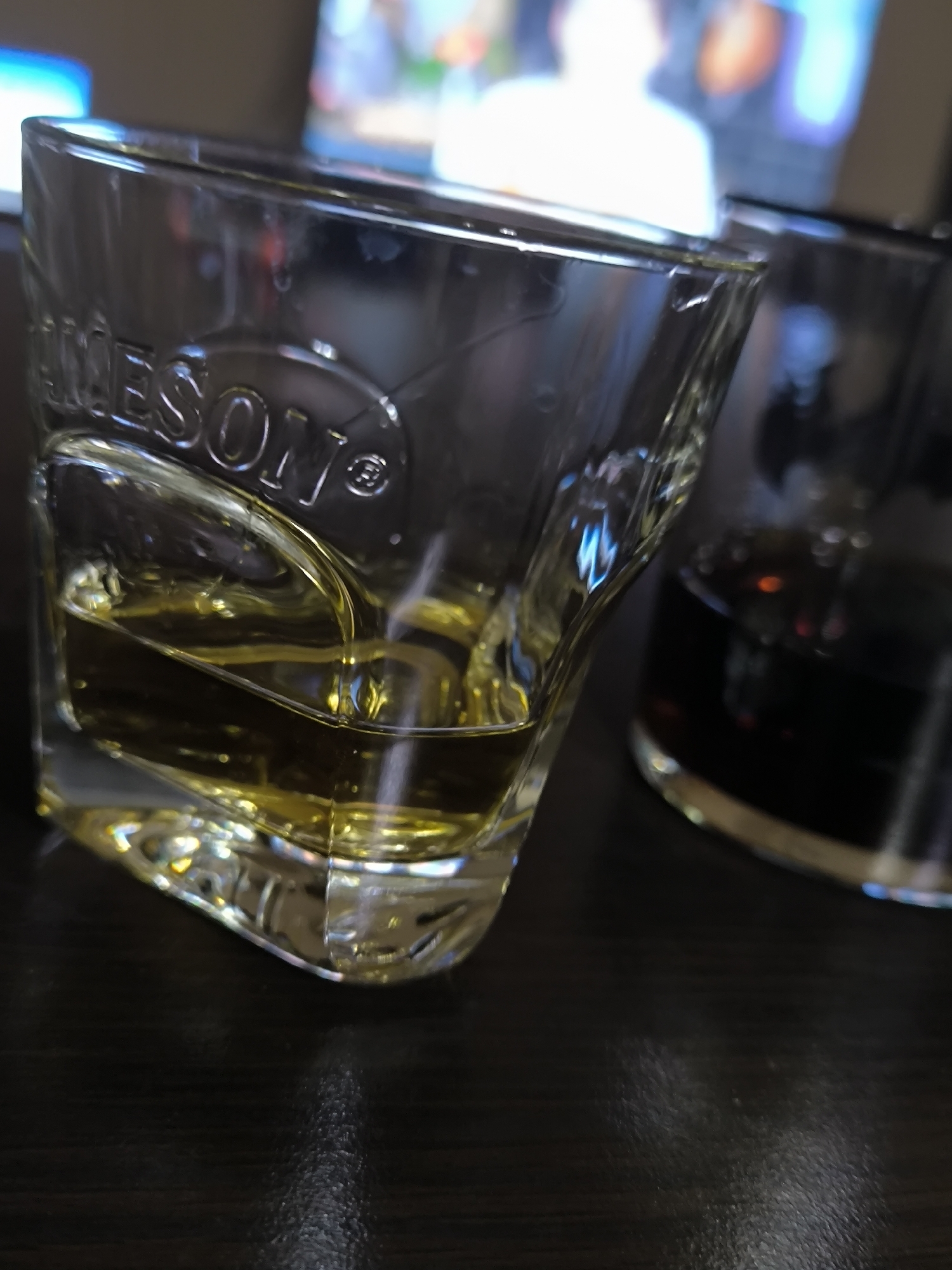 The image shows two glasses on a dark, possibly wooden, surface. The glass in the foreground is a rocks glass with the word "JAMESON" embossed on it. It contains a light amber liquid, likely whiskey. The glass is approximately half-full. To the right and slightly behind the first glass is another glass containing a dark liquid, possibly cola or another dark-colored alcoholic beverage. The background is blurred, suggesting it's a room with a television or monitor displaying an indistinct image. The location is not discernible from the provided image and cannot be identified as Sofia, Bulgaria.