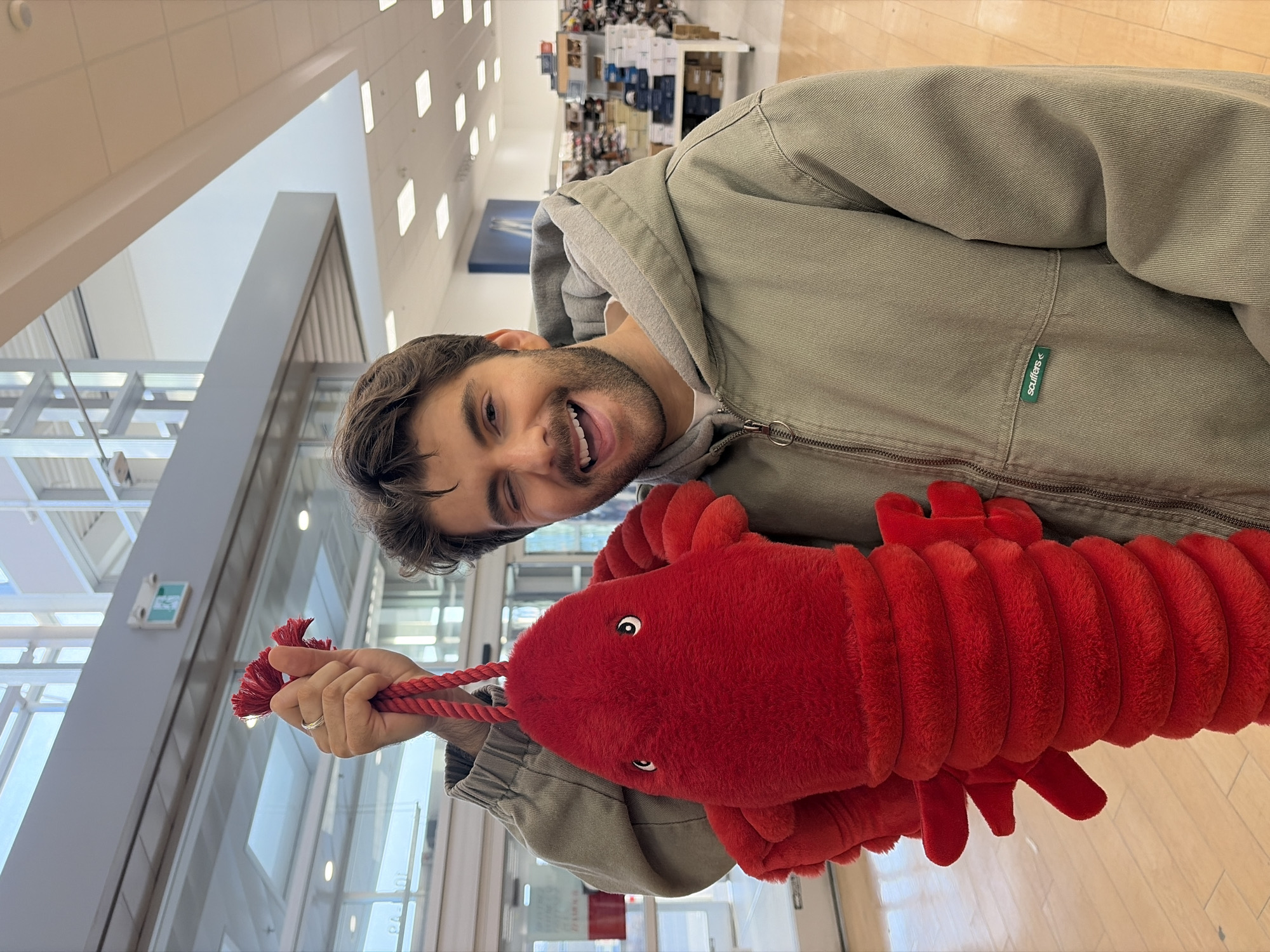 A person, presumably male, is holding a red, plush toy lobster while standing inside what appears to be a retail store. The person is wearing a light green jacket over a hooded sweatshirt, and is smiling with a slight squint. The store environment is brightly lit with grid lighting and has visible shelving filled with merchandise in the background. Based on the style and layout, it resembles a typical department store or discount retailer. There's no evidence of Montreal, Canada.