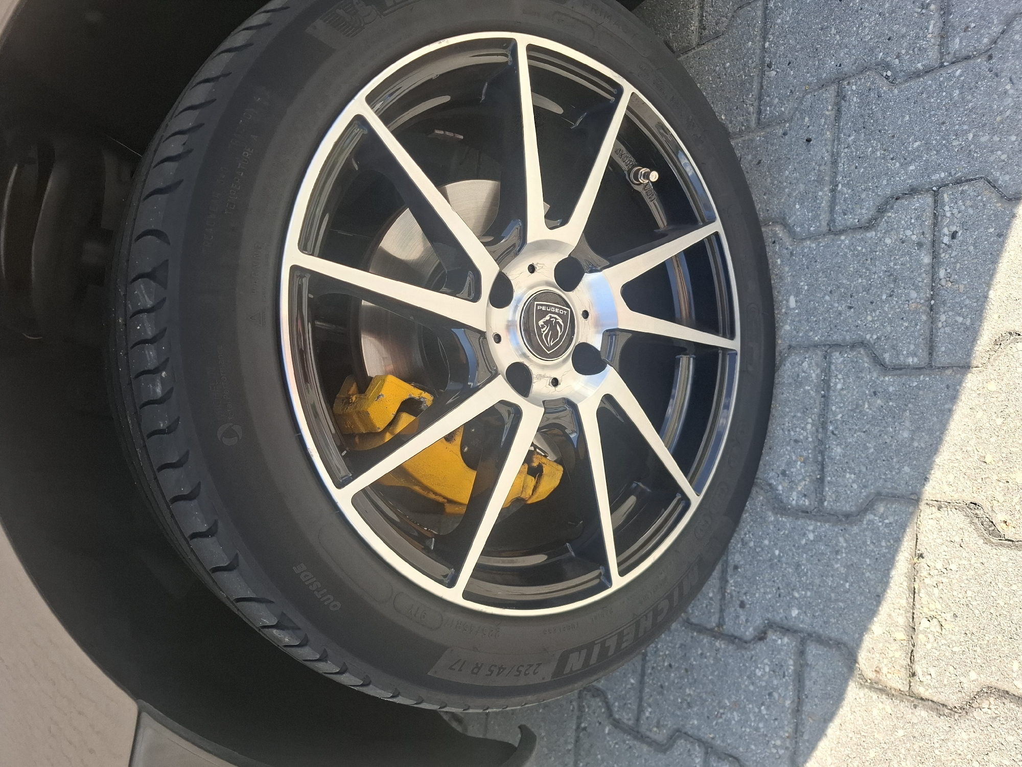 A close-up, eye-level shot shows the front passenger-side wheel of a car parked on a paved walkway made of interlocking grey concrete blocks. The car's exterior is a muted brown. The wheel features a silver and black alloy rim with five sets of double spokes. The brake caliper is painted a bright yellow, and the tire is a Michelin brand, with "225/45 R 17" visible on its sidewall. The image does not contain any people. The scene is devoid of any identifying landmarks that would indicate a specific location in Bratislava, Slovakia.