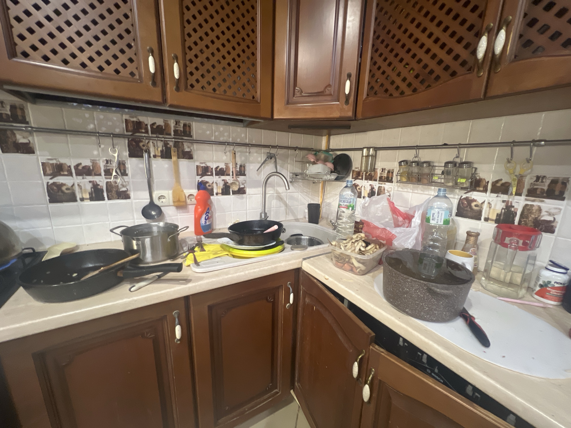 The image depicts a cluttered kitchen counter in what appears to be a residential setting. The scene is dominated by dark wooden cabinets and a light-colored countertop. On the counter, there are various kitchen items, including pots, pans, dishes, bottles of water, and cleaning supplies. A sink is visible with a faucet. The backsplash is tiled with a decorative pattern. The overall impression is of a lived-in kitchen that may be in the process of being used or cleaned. There are no people visible in the image. The location is identified as Трещиха, Russia.