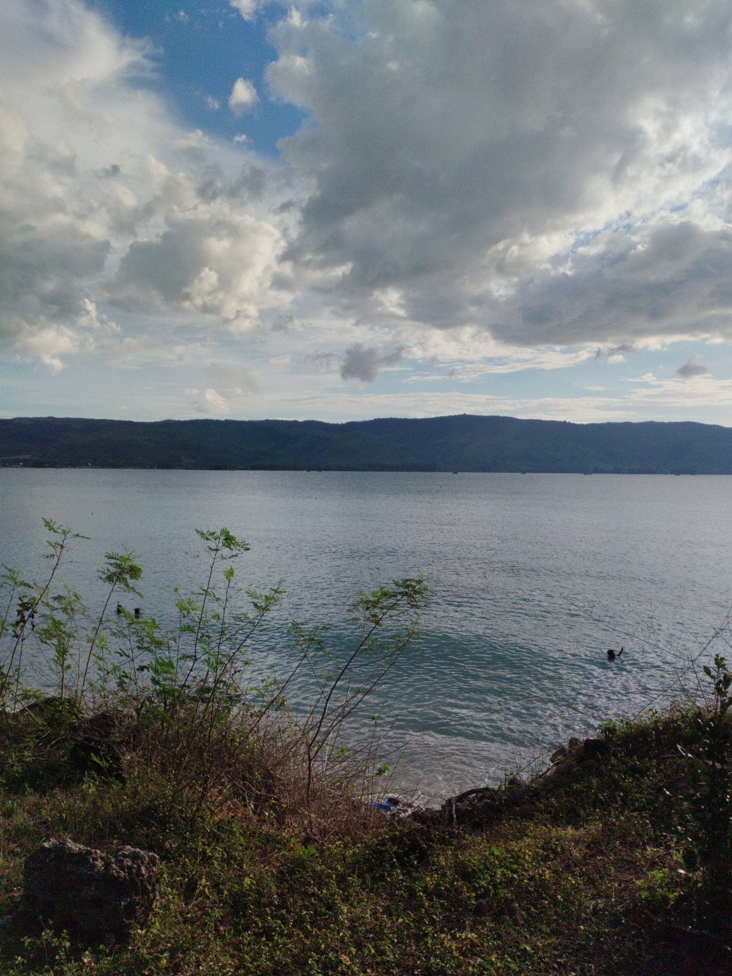 The scene depicts a coastal view in Lamreh, Indonesia, under a cloudy sky. A body of water fills the midground, reflecting the light. The far shore is marked by a dark, elevated landmass. In the foreground, there is vegetation, including shrubs and small trees. A person is swimming in the water near the shoreline.