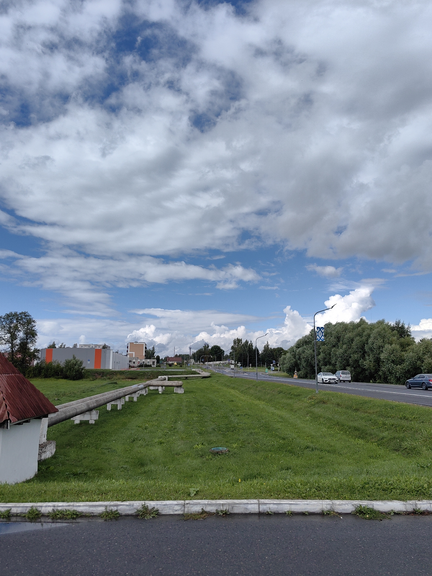The image depicts a scene along a road in Myadzyel, Belarus. In the foreground, a paved road with a light-colored curb is visible, with some grass and small plants growing along the edge. To the left of the road, there is a grassy embankment leading up to a small white building with a red-tiled roof. Two large, insulated pipes run horizontally across the grassy area from the building, supported by concrete pillars. Further into the distance, a multi-lane road stretches out, with several vehicles, including a white car and a silver SUV, driving on it. On the right side of the road, a dense line of green trees forms a backdrop. Beyond the road and trees, several buildings with modern architectural styles are visible, including one with orange and grey sections. The sky is a vibrant blue with scattered white and grey clouds. The overall scene suggests an industrial or semi-urban environment on a cloudy day.