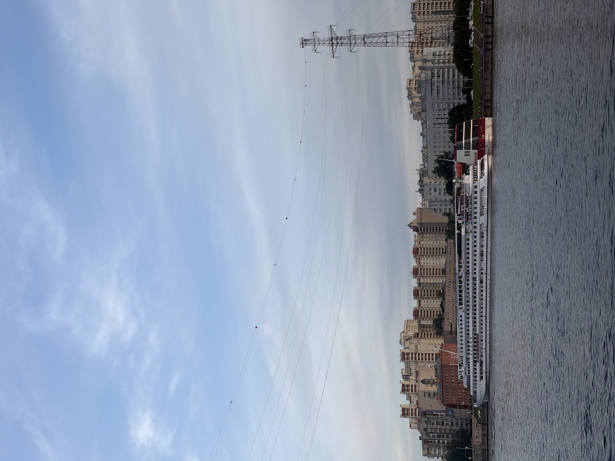 The image presents a wide outdoor scene along a riverfront in an urban area within Russia. The sky occupies the top half, displaying a light blue hue with scattered, wispy white clouds. Multiple parallel power lines stretch across the upper mid-ground, featuring alternating white and orange spherical marker balls for visibility. On the far right, a tall, metal lattice-work electricity transmission tower stands against the sky, extending above the city skyline.
The lower half of the image features a wide body of water, likely a river, its surface rippled and dark. Along the far bank, a concrete or stone embankment is visible. Docked parallel to this embankment are two large multi-deck vessels. The prominent vessel on the left is a white river cruise ship with "ЛЕНИН" (LENIN) inscribed in red capital letters on its side. To its right, a second, smaller multi-deck boat with a distinctive red and white superstructure is also docked.
Behind the embankment and boats, a dense cluster of multi-story residential buildings forms the city backdrop. These structures exhibit architectural styles common in Russian cities, primarily light-colored facades with some brown or red roof sections. A narrow strip of land with sparse trees and a visible roadway with a few parked cars runs between the embankment and the buildings. The scene appears devoid of discernible human activity, presenting a static urban landscape under natural daylight.