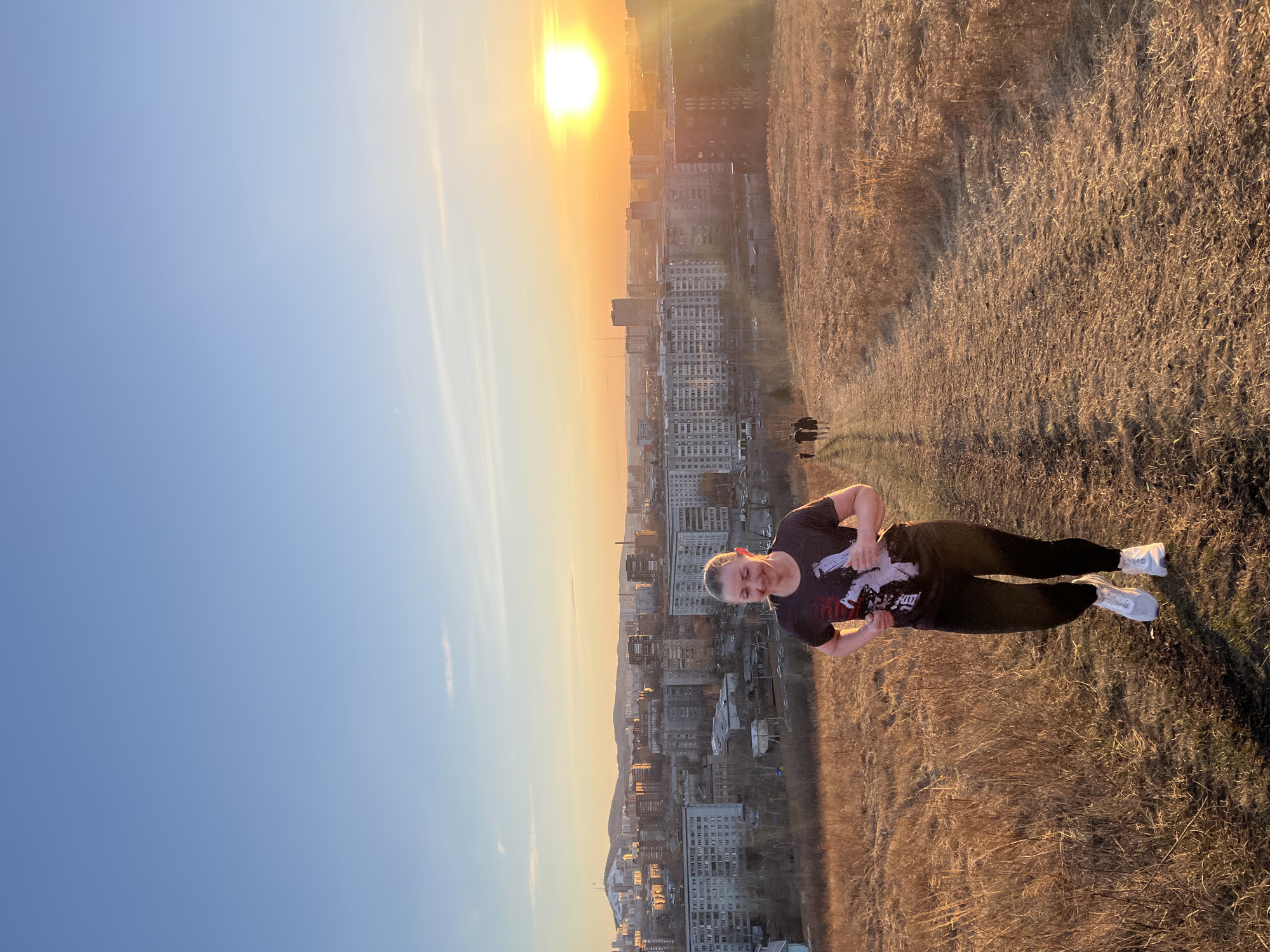 A portrait-oriented photograph captures a woman standing on a dry, grassy hillside, overlooking a sprawling city under a radiant sunset or sunrise. The woman, positioned towards the right of the frame, has short, light-colored hair and is smiling faintly towards the camera. She is dressed in a dark t-shirt with a white and possibly red graphic design on the front, dark pants or leggings, and white athletic shoes. Her hands are clasped in front of her.
The foreground and midground consist of a sloping hillside covered in dry, golden-brown grass, suggesting a seasonal transition or dry climate. Below the hillside, a vast urban landscape stretches out, filled with numerous multi-story buildings, typical of residential or commercial areas in a large city like Krasnoyarsk, Russia.
The sky is a prominent feature, transitioning from a clear light blue overhead to a vivid orange and golden hue near the horizon where the sun is brightly visible and low. This strong light casts a warm glow over the entire scene and indicates either dawn or dusk. Long shadows are visible on the hillside, further emphasizing the low angle of the sun. The overall impression is one of peaceful enjoyment of a scenic overlook, contrasting natural terrain with an urban environment under a dramatic sky. No discernible text is visible on the woman's clothing or within the scene.