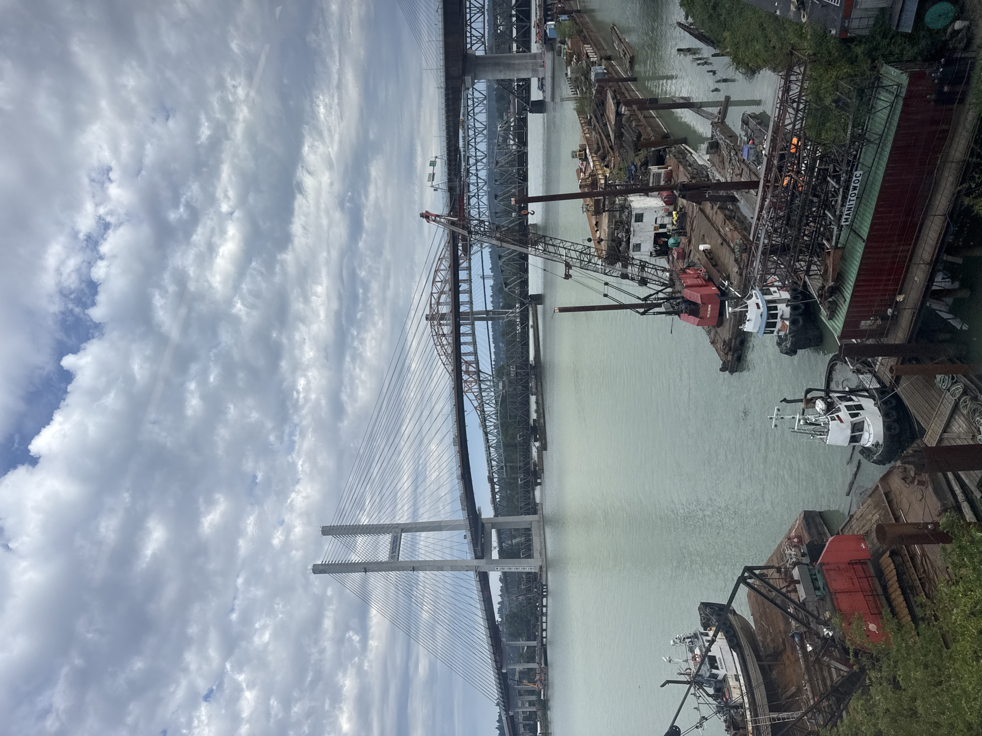 The image depicts a wide river with several bridges spanning it, indicative of an urban or industrial waterfront. In the foreground, barges are docked, some laden with construction materials and equipment, including cranes and shipping containers. Several smaller boats, possibly tugboats or service vessels, are also present. The water appears somewhat murky.
In the background, multiple bridges are visible. The most prominent is a large, modern cable-stayed bridge with tall, concrete towers. Behind it, an older, truss bridge is also present. Further in the distance, a third, darker bridge is visible. The sky is overcast with a mix of clouds and some patches of blue. The banks of the river show signs of development, with some vegetation and structures.
Based on the presence of these specific bridges and the general industrial and port-like atmosphere, this scene is highly likely located in New Westminster, Canada, specifically along the Fraser River. The bridges visible are characteristic of the Port Mann Bridge (cable-stayed), the Pattullo Bridge (truss), and potentially the Canadian National Railway Bridge in the vicinity. The activity on the barges suggests ongoing construction or maintenance work in the area. There are no discernible individuals in the image.