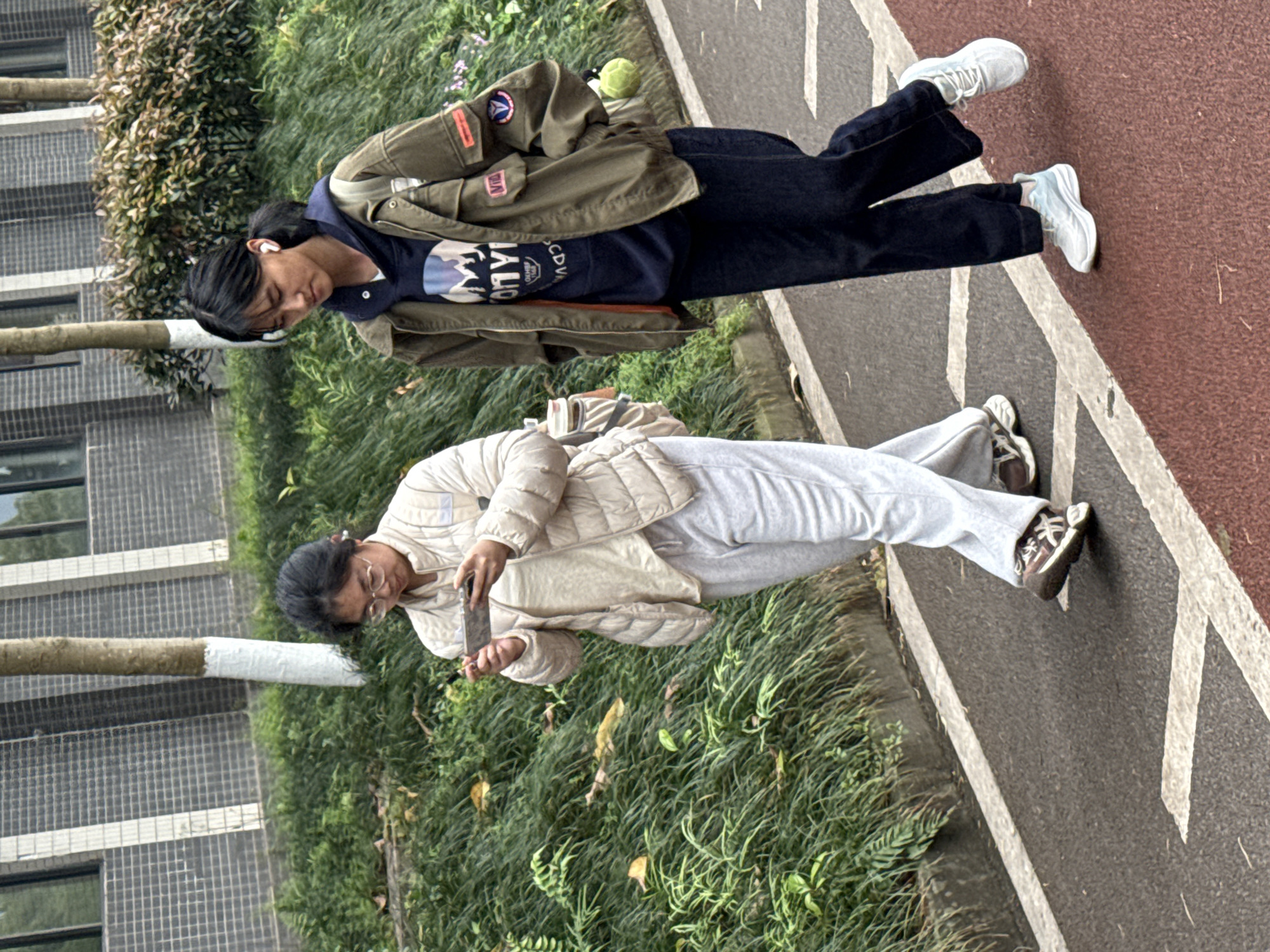 Two individuals stand on a paved walkway adjacent to grass and trees. The walkway is marked with white lines, with a red painted section alongside. A building is visible in the background. The first person is wearing a green jacket, a navy blue shirt, and dark pants, with white sneakers. The second person is wearing a light beige jacket and light grey pants, with tan-colored shoes. The second person is holding a phone and appears to be taking a picture. The location is urban. The city is Jiangjin, China.