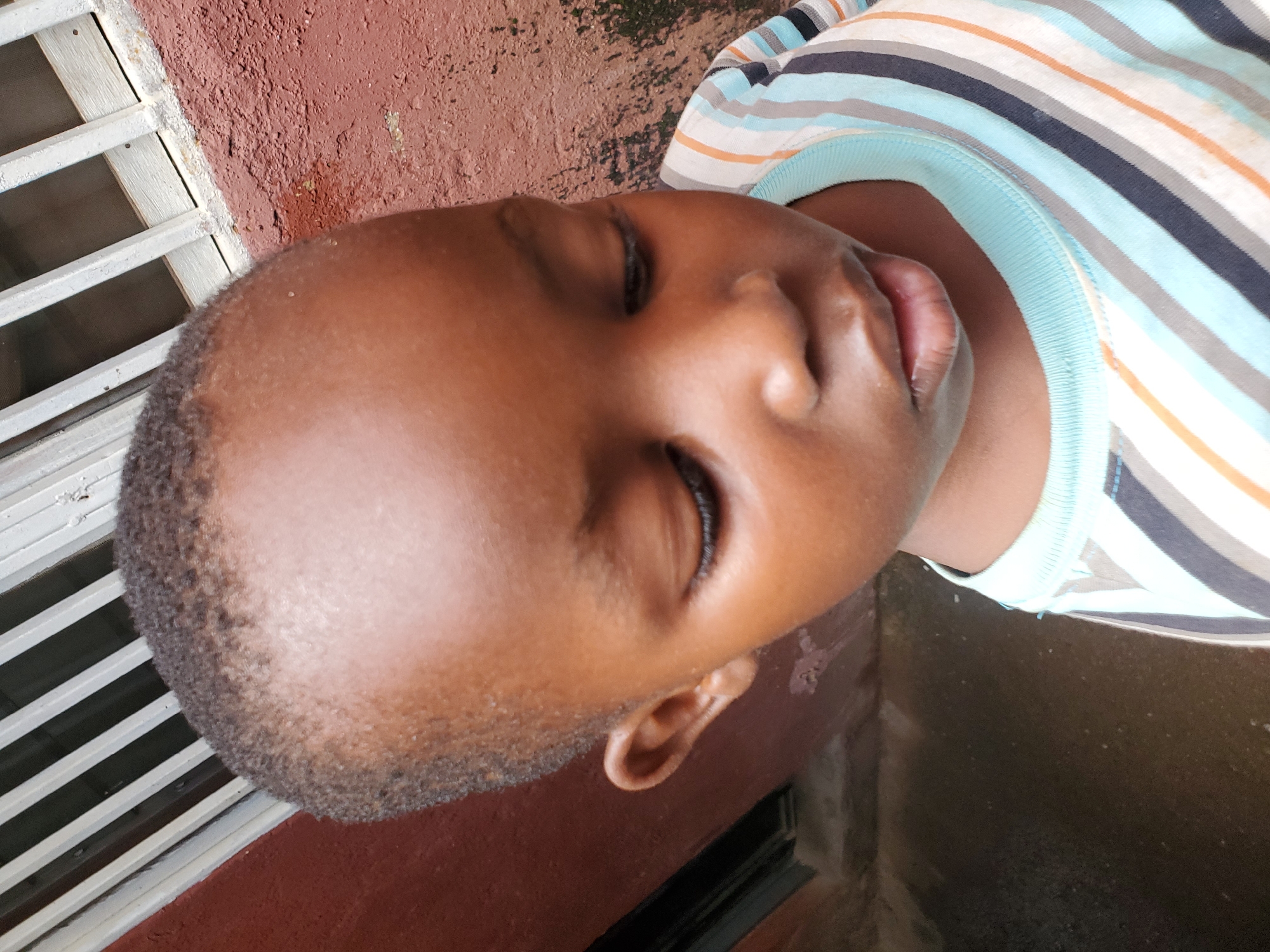 The media file shows a close-up, rotated shot of a young Black child's face, from the nose upwards, as they are lying down. The child is wearing a horizontally striped t-shirt with blue, white, orange, and dark gray stripes, and a light blue neckline. Their eyes are closed, and their mouth is slightly open, suggesting they are resting or asleep. The child's skin tone is a medium brown.
In the background, to the left, is a portion of a white grated window or barrier against a textured, reddish-brown wall. To the right of the child's head, the ground appears wet and dark, possibly indicating recent rain or a damp surface. The lighting suggests it could be daytime, possibly mid-morning or afternoon, as there are bright highlights on the child's skin and the wall. The scene seems to be outdoors, given the wall and ground textures and the natural light. No text is visible in the image. The overall impression is one of peaceful repose in a simple outdoor setting.