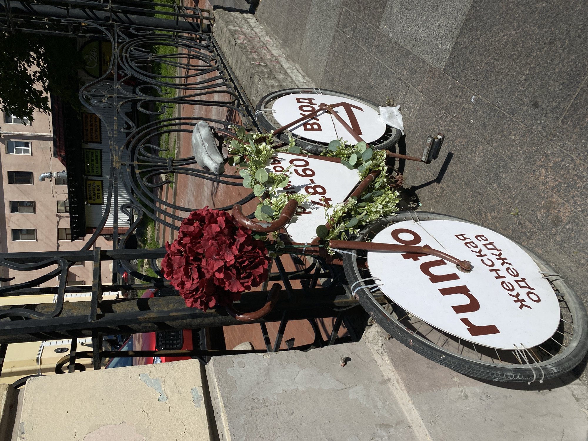 An outdoor scene in Saint Petersburg, Russia, captured on a bright, sunny day, shows a repurposed bicycle functioning as a commercial sign, leaning against an ornate black wrought-iron fence. The bicycle, painted brown, is decorated with a large, vibrant red artificial flower arrangement near the handlebars and a green leafy garland around its frame.
The wheels of the bicycle are fitted with white circular signs. The front wheel sign displays "Вход" (Russian for "Entrance") in brown lettering, indicating an entry point. The rear wheel sign features "runto женская одежда" (Russian for "runto women's clothing"), advertising a retail business. A rectangular white sign attached to the bicycle frame shows the number "48-606," likely part of a phone number.
Behind the fence, a light-colored building is visible. On its facade, multiple signs are present. One green sign, partially obscured, shows "24ч" (24 hours), suggesting an all-day service. Another prominent white sign lists a menu in Russian: "ШАВЕРМА, ФАЛАФЕЛЬ, ХОТ-ДОГ," along with "КОМБО-ОБЕД: ПЕРВОЕ ВТОРОЕ, САЛАТ НАПИТОК," and "КРЕВЕТКИ, НАГГЕТСЫ, КАРТОШКА ФРИ," indicating a fast-food establishment or cafe nearby.
The ground is a paved sidewalk made of speckled, dark-colored tiles. The overall impression is an urban setting, utilizing a creative display for local businesses.