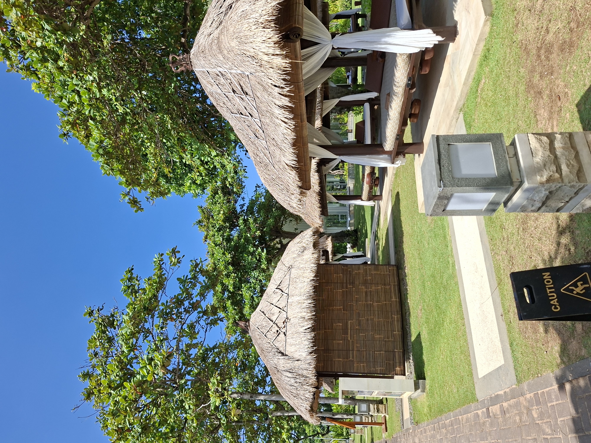 The image depicts two thatch-roofed gazebos, characteristic of a tropical resort, in Kuta Selatan, Indonesia. The gazebos are positioned alongside a paved pathway and a lawn. One gazebo is open, with white draped curtains and lounge seating. The other appears enclosed with woven panels. The sky is clear and blue, visible behind the foliage of a tree partially framing the scene. A "Caution: Wet Floor" sign stands near the walkway. A stone and glass-encased light fixture is also present along the pathway. No people are visible.