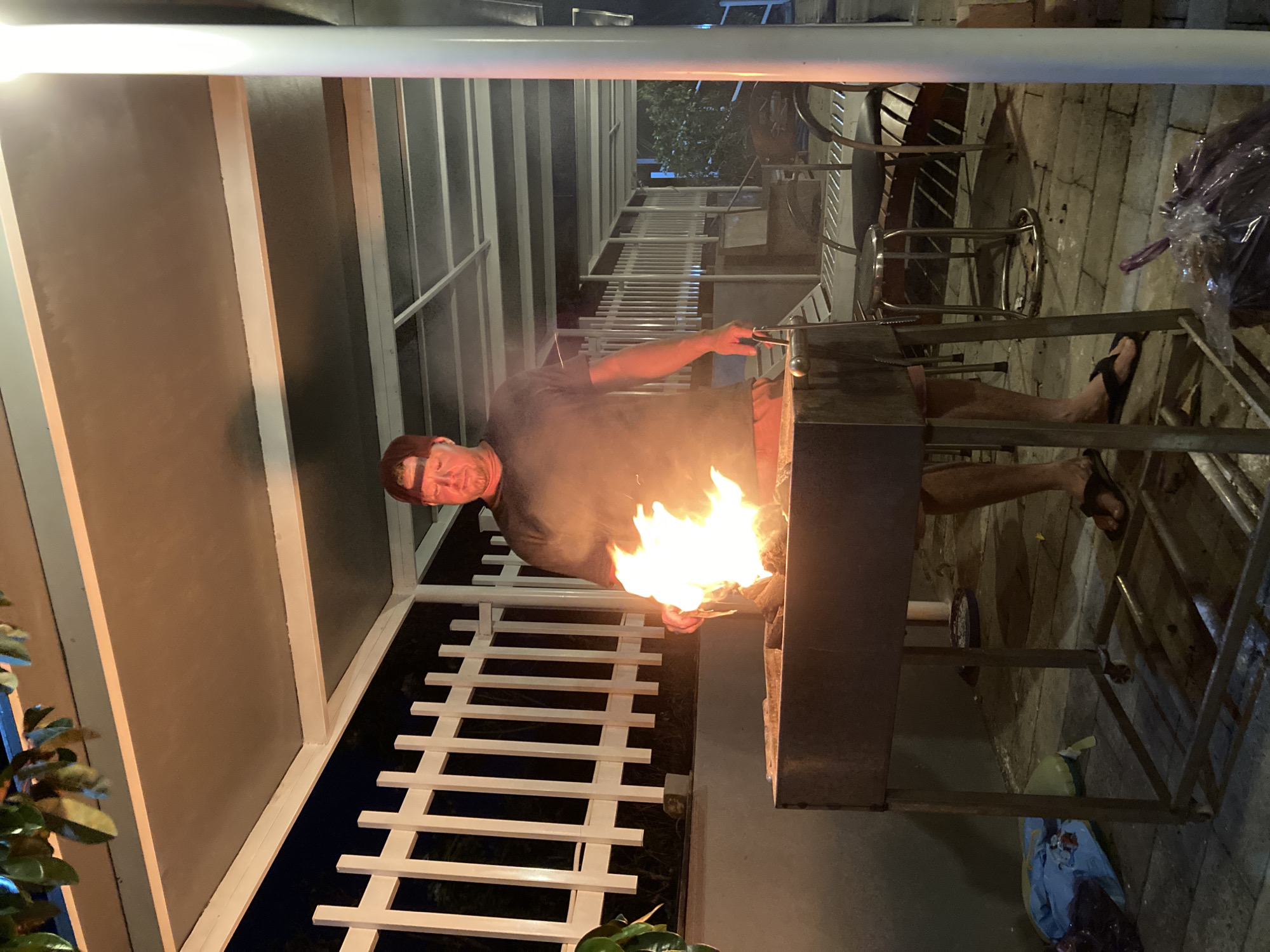 A man is standing next to a barbecue grill with a large flame inside. He is wearing a dark-colored t-shirt, and possibly shorts, and is wearing sandals or flip-flops. The grill is on a metal stand and is located on a tiled patio or walkway. There is a white railing and wall structure in the background. Overhead lighting is visible. The location is Mũi Né, Vietnam.
