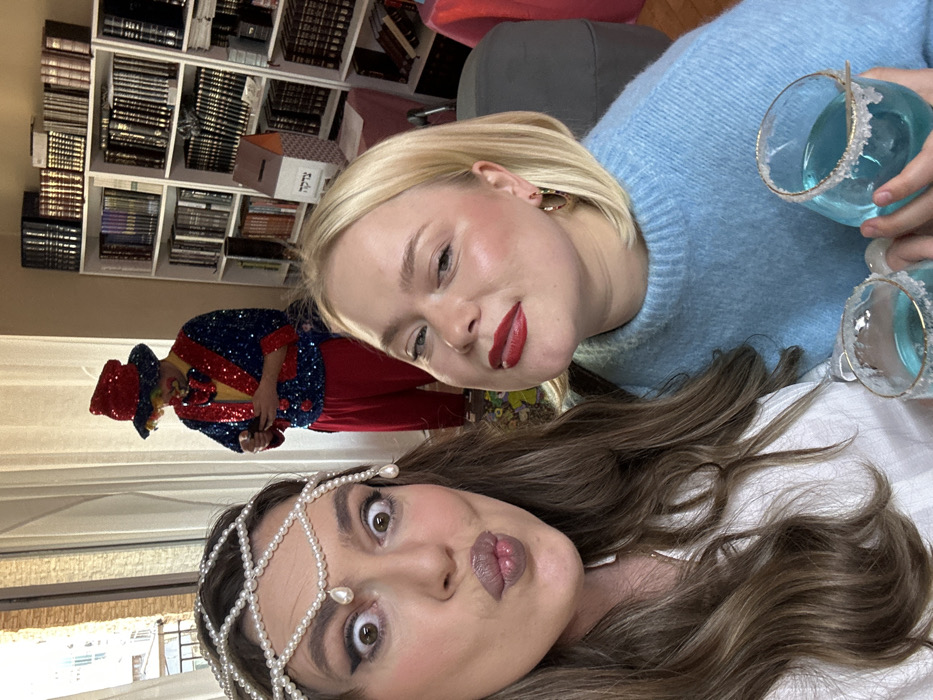 This indoor photograph depicts a festive gathering featuring three individuals. In the foreground, a woman with long brown hair wears a pearl-adorned headpiece and a white top, making a wide-eyed expression with puckered lips directly at the camera. To her right, a second woman with a blonde bob haircut and red lipstick, dressed in a light blue sweater, holds a glass containing a blue beverage with a sugared rim; she is also looking towards the camera. In the background, positioned between the two foreground subjects and a set of bookshelves, a third individual is dressed in a full clown costume consisting of a red and blue sequined jacket, red pants, a yellow shirt, and a red sequined top hat with a colorful wig; this person is holding and looking at a mobile phone. The background on the right features two white bookshelves filled with numerous books, primarily with dark bindings. On a table in front of the right bookshelf, a light brown cardboard box marked with the Hebrew word "צדקה" (Tzedakah, meaning charity) is visible. To the left of the clown, a large window or balcony door reveals an exterior view of a building, indicative of an urban setting. The overall context suggests a celebratory event, likely a Purim party, within a Jewish community space in the city of Tbilisi, Georgia.