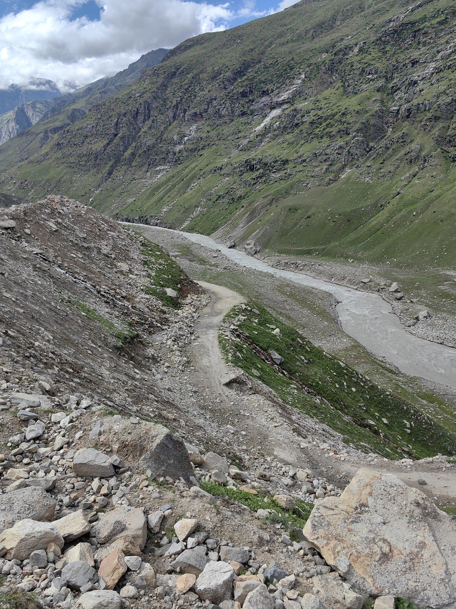 A vertical, eye-level, medium shot captures a rocky, mountainous landscape in India under a partly cloudy sky. In the foreground, a steep, rocky slope is covered in loose stones and pebbles. A narrow, unpaved dirt path winds its way down the slope, bordered by more rocks and sparse green vegetation. To the right and extending into the background, a wide, grey river flows through a valley. The opposite side of the valley is a steep, green mountainside covered in grass and scattered rocks, with a visible streak of what appears to be a dry waterfall or water runoff. The mountains in the distance are shrouded in clouds, adding to the rugged, natural beauty of the scene. The lighting suggests it is daytime.