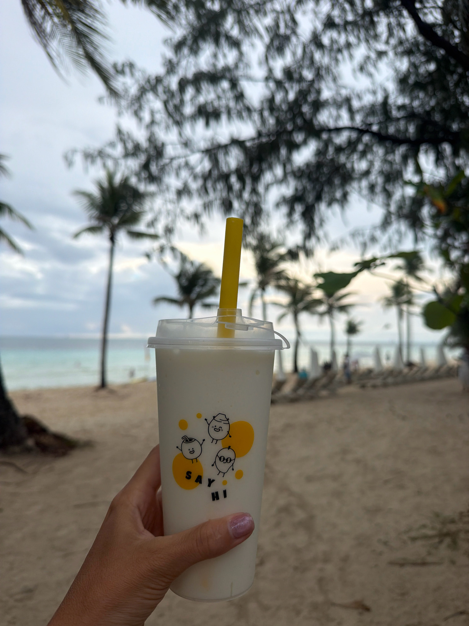 A person's hand, with fingernails painted pink, holds a white plastic cup containing a light-colored beverage and a yellow straw. The cup has a transparent plastic lid with a hole for the straw. The cup is decorated with a yellow heart and three cartoon-like characters, along with the text "SAY HI" in black lettering. The background is a beach scene in Malay, Philippines, with a sandy shore, palm trees, lounge chairs, and a body of water under a cloudy sky.