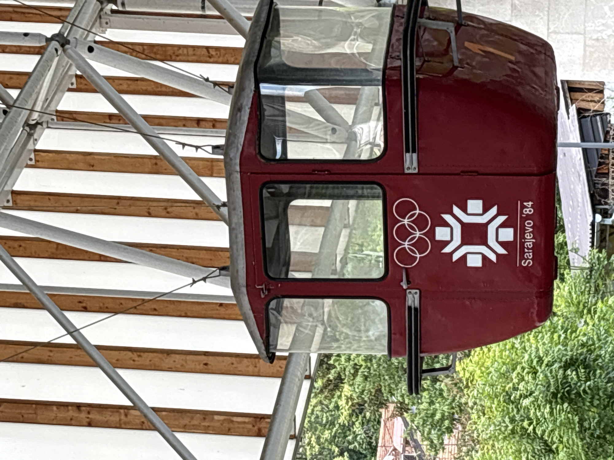 A slightly tilted, close-up shot shows a red gondola cabin displaying the Sarajevo '84 Olympics logo. The cabin has multiple windows, revealing portions of the steel framework. The environment includes elements of foliage and structural supports.