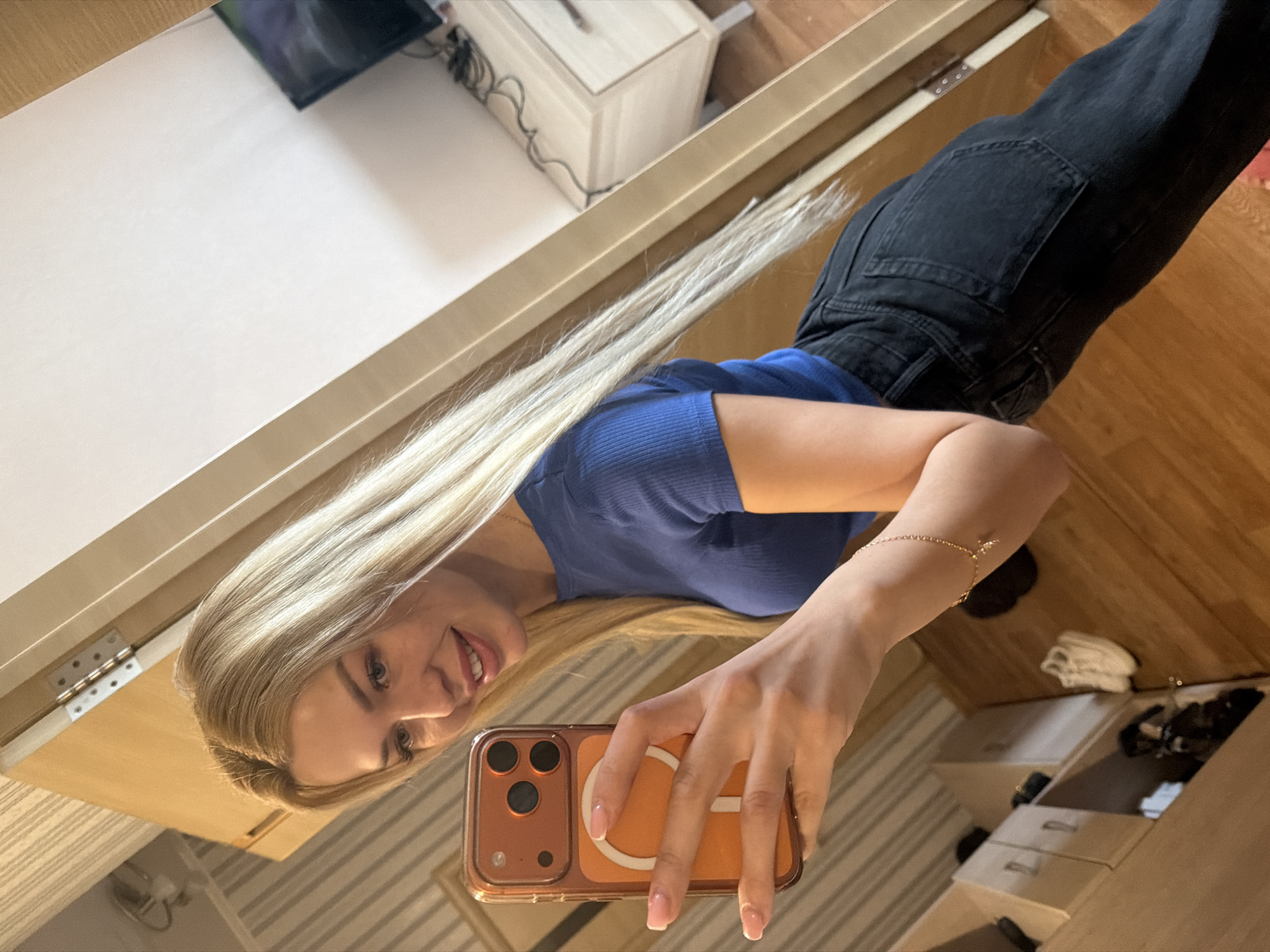 A young blonde woman in a blue t-shirt and black jeans is taking a mirror selfie in what appears to be a home setting in Kyiv, Ukraine. She is holding a phone with an orange case and is wearing a delicate gold bracelet. The background shows a wooden floor, a closet with shelves containing shoes and other items, and a dresser with a television screen visible in the reflection of a large mirror. The woman is smiling and looking at her phone.