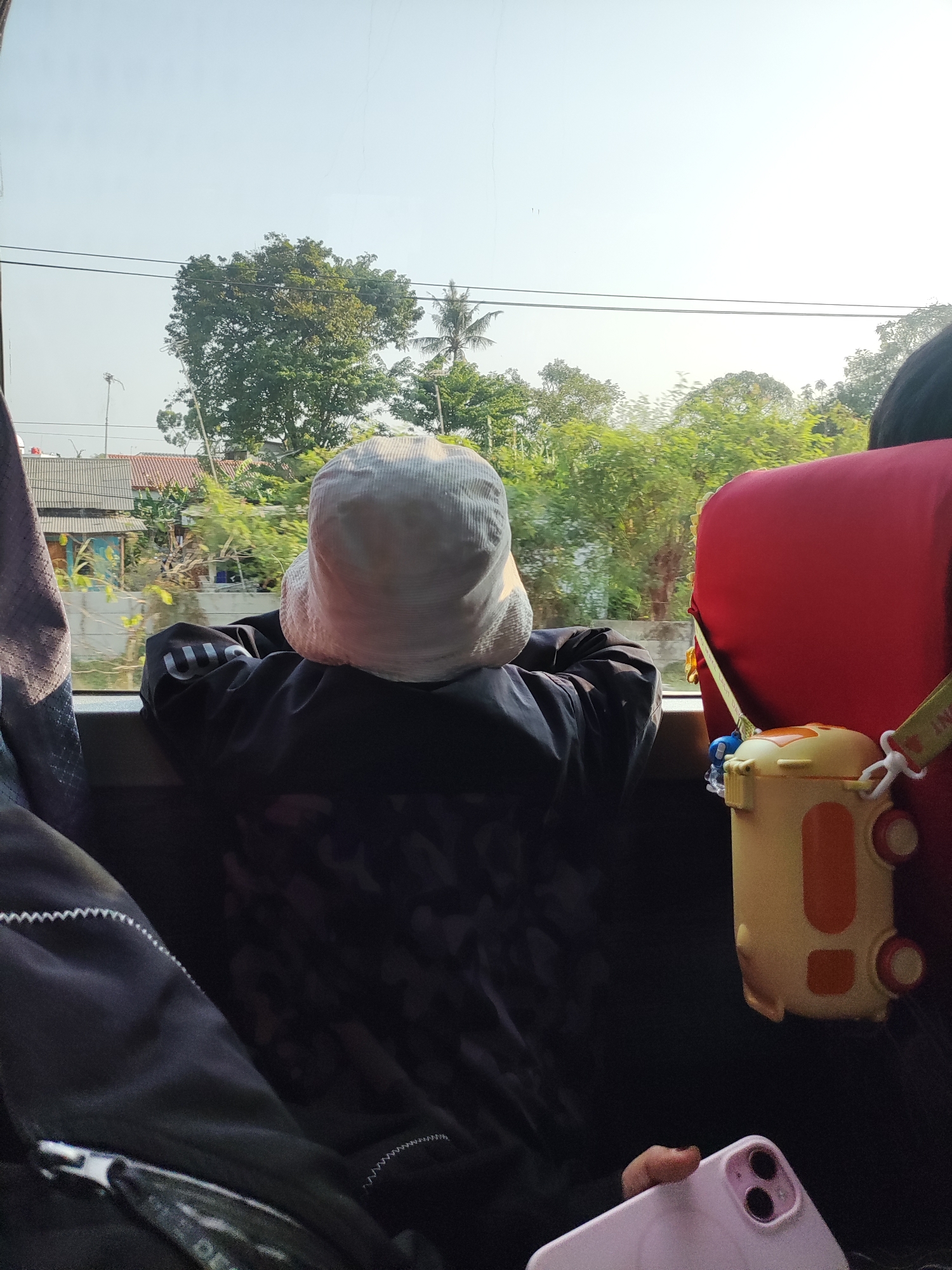 The image shows a rear view of a person wearing a white bucket hat and a dark jacket with a floral pattern, leaning on a window. The person is likely a child, based on their size. The scene appears to be inside a vehicle, possibly a bus or car, as indicated by the window and the seats visible. Outside the window, trees, houses with red tiled roofs, and power lines are visible, suggesting a semi-urban or rural environment. The sky is clear and blue. To the right of the person, a red seat and a yellow container with a strap are visible. In the foreground, a pink smartphone is on a dark surface. The overall lighting suggests it is daytime. The mention of Karawang, Indonesia, places this scene in a specific geographical context.