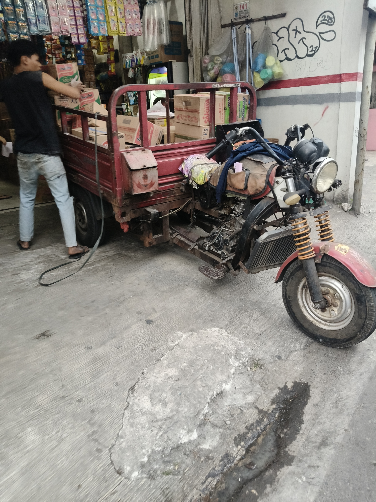 A young male is loading boxes into the back of a three-wheeled vehicle that is parked on a concrete surface. The vehicle is red and has a motorcycle-like front end. The male is wearing a black t-shirt and light blue jeans. He is standing to the left of the vehicle and is reaching into the cargo area. The cargo area is filled with various boxes. Behind the vehicle is a store with shelves displaying products. The store appears to be located in South Tangerang, Indonesia, as indicated by the presence of local products and signage typical of Indonesian markets. The scene is outdoors, with a wall visible in the background that has graffiti on it. The concrete ground shows some signs of wear and tear, including a dark stain that appears to be oil or grease.