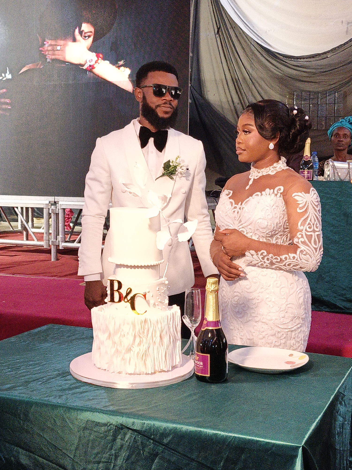 In Fagge D2, Nigeria, a wedding scene unfolds indoors. A man in a white suit, bow tie, and sunglasses stands beside a woman in a long-sleeved white lace dress. They are positioned behind a table draped in a teal fabric. On the table is a three-tiered white wedding cake with gold lettering "B & C", a bottle of sparkling wine, a plate, and two champagne flutes. Behind them, there's a screen displaying a partial image of another couple. A woman wearing a head covering stands behind the table in the background, near what appears to be a silver purse. The setting seems to be a banquet hall or reception area.