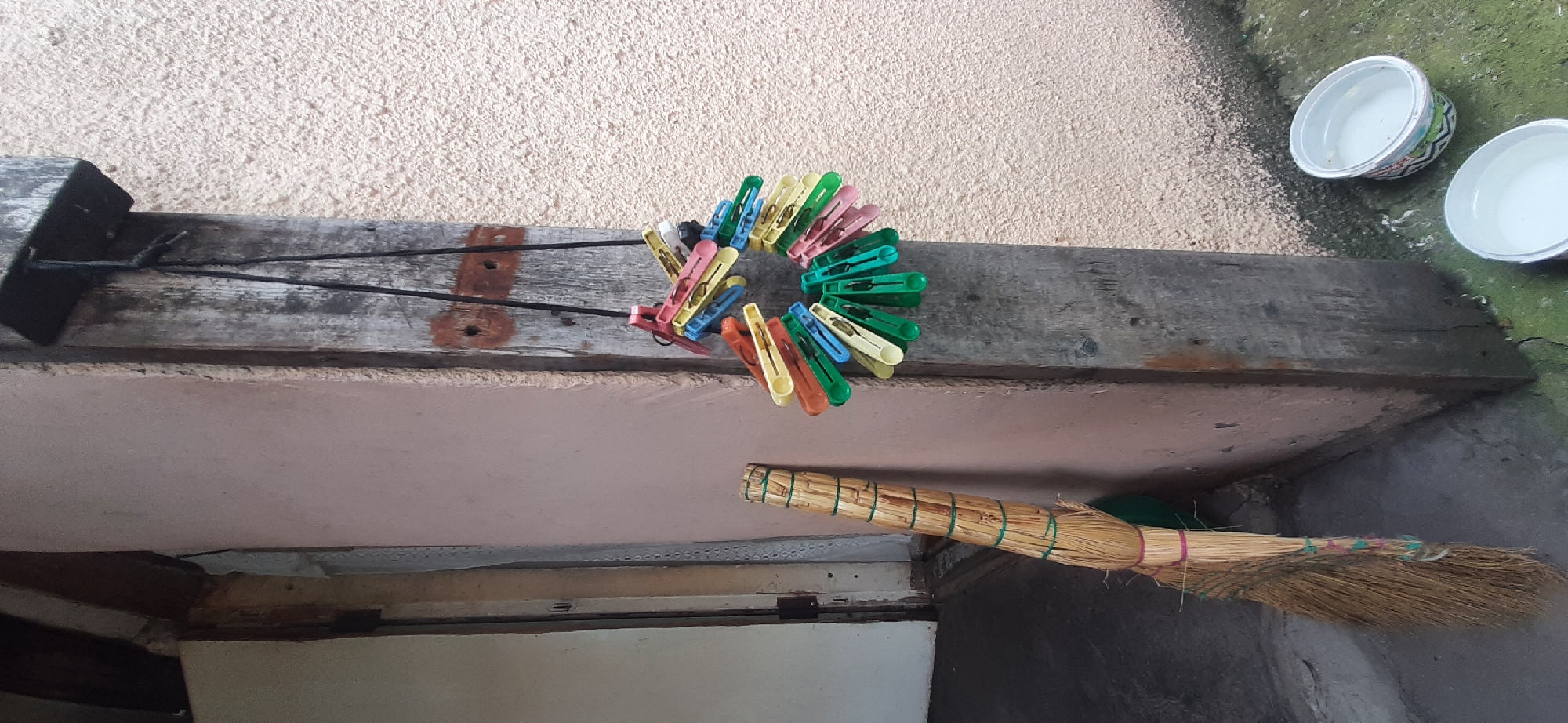 This is a close-up, eye-level shot of an outdoor scene, possibly a patio or balcony, in Yessentukskaya, Russia. The main subjects are a collection of colorful clothespins arranged in a circular pattern on a weathered wooden beam, and a broom made of natural straw, also resting on a wooden surface.
The clothespins, in various shades of green, blue, yellow, pink, and orange, are clipped together and form a spiral or flower-like shape. They appear to be attached to two thin black wires that are strung between a dark, rectangular object on the left and the wooden beam. The wooden beam itself shows signs of age, with visible grain and some reddish staining.
Below the beam, a light pink painted surface is visible, and further down, a section of what looks like a window frame or cabinet is present. The broom, positioned horizontally to the right, is tied with green and pink bands around its straw bristles.
In the upper right corner of the image, two white plastic bowls are visible. One contains a small amount of liquid, and both appear to be resting on a rough, greenish-gray surface that could be a wall or the ground.
The lighting suggests it is daytime, with diffuse light, possibly indicating an overcast sky. The overall impression is of a functional, outdoor area, perhaps a laundry drying area, with everyday items stored or left out. There is no visible text in the image.