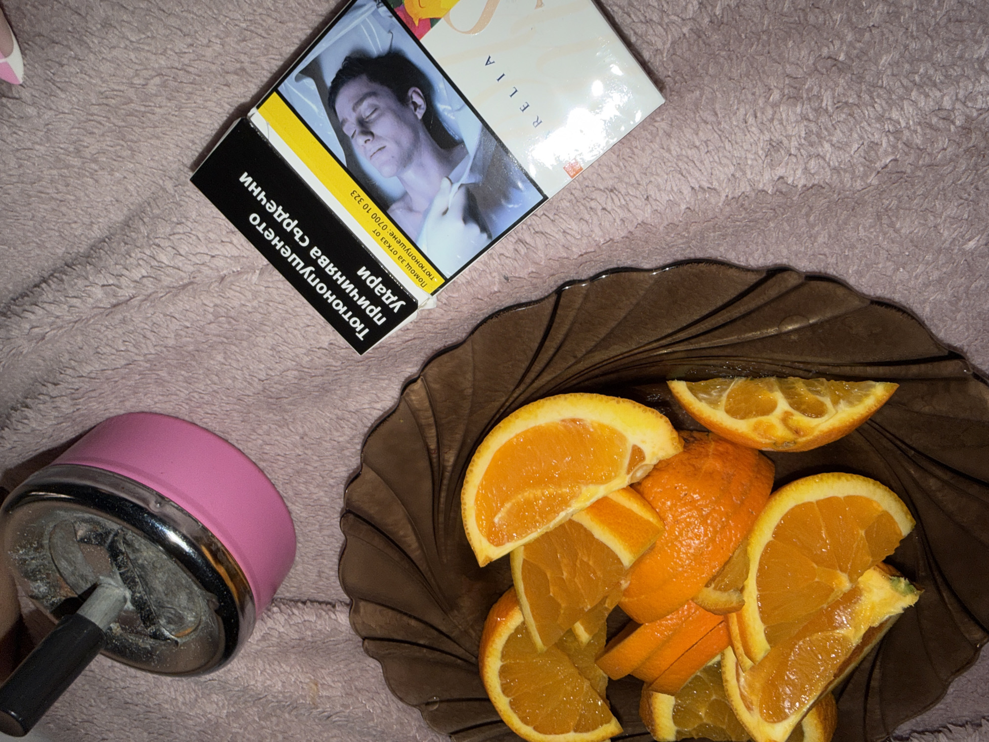 The scene depicts a close-up shot on what appears to be a soft, textured surface. A dark, translucent bowl contains a pile of orange slices and sections. Next to the bowl sits a pink and silver-colored ashtray containing what appears to be cigarette residue. A packet of cigarettes, branded "RELIA", is placed near the bowl. The image on the cigarette packet features a man lying down. The packaging includes warning labels and information in Cyrillic script. The location is not visually discernible and cannot be determined based solely on the objects within the frame.
