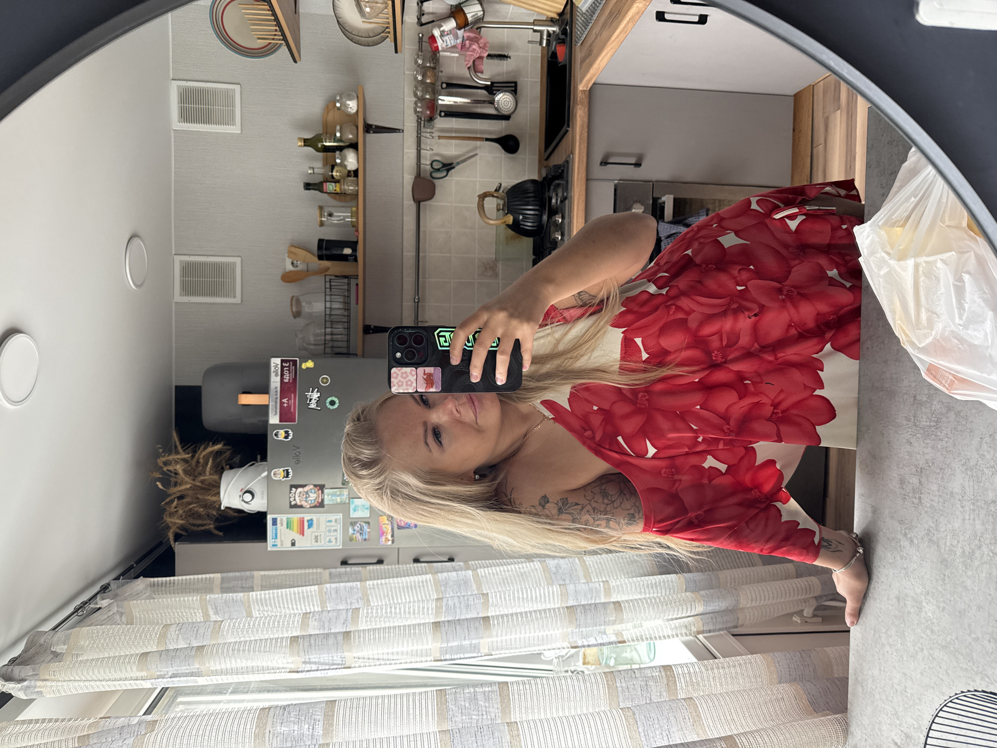 A woman is standing in front of a round mirror, taking a selfie with her phone. She is wearing a red and white floral dress. Her blonde hair is long and styled to one side, and she has tattoos on her arm. The background shows a kitchen with various items on shelves and hanging on the wall. A refrigerator covered in magnets and stickers is visible, and there are curtains in front of a window. A plastic bag is visible on the countertop in the mirror's reflection.