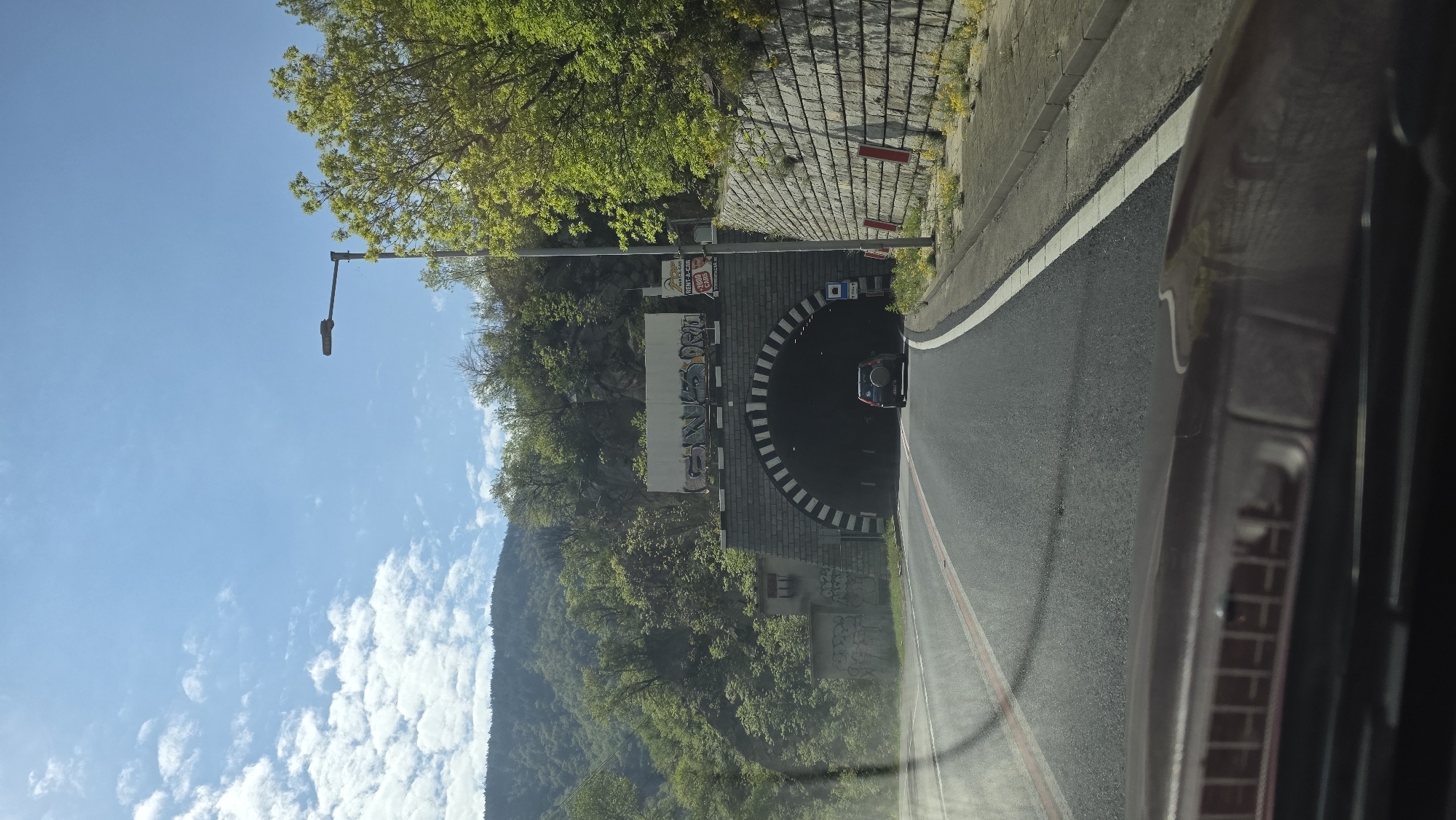 The image captures the view from inside a car, seemingly approaching the entrance of a tunnel located in Stara Kresna, Bulgaria. The road leading to the tunnel is paved and marked with lane dividers. The tunnel's entrance is characterized by alternating black and white stripes forming an arch. To the left of the tunnel entrance, a mountain covered with green trees and vegetation is visible. The sky above is blue with scattered white clouds. A street lamp stands nearby. A car is emerging from the tunnel. The right side of the image is dominated by the interior of the vehicle from which the image is taken.