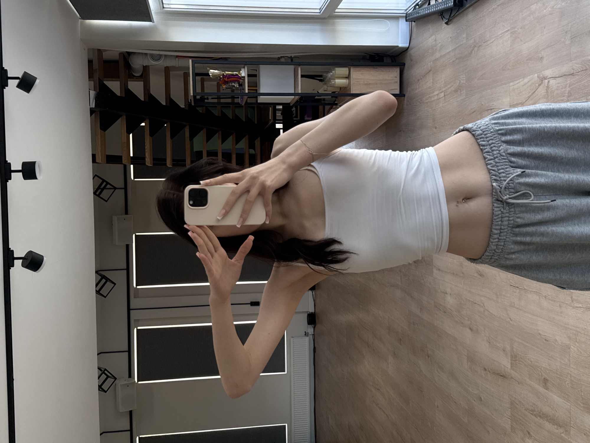 The media file shows a person taking a mirror selfie in what appears to be a modern apartment or studio in Pokrovskoe, Russia. The person is wearing a white tank top and grey sweatpants. They are holding a smartphone with a light pink case in their right hand, with their left hand slightly raised. Their reflection is visible in a large mirror that covers a significant portion of the wall.
In the background, seen through the mirror, is a staircase with black railings and wooden steps, suggesting an interior space with multiple levels. There are shelves mounted on the wall above the staircase, displaying various objects, including what look like decorative items and bottles. The overall aesthetic suggests a contemporary design.
The lighting in the room appears to be artificial, with track lighting visible on the left side of the frame and light strips integrated into the wall panels behind the person. The floor is made of light-colored wood planks.
The image is oriented horizontally, and the person's body is mostly visible from the torso down. The focus is on the person and their outfit, as well as the surrounding interior elements. There is no visible text in the image. The time of day is not discernible, and the weather is irrelevant to the interior setting. The overall mood is casual and relaxed, typical of a personal selfie.