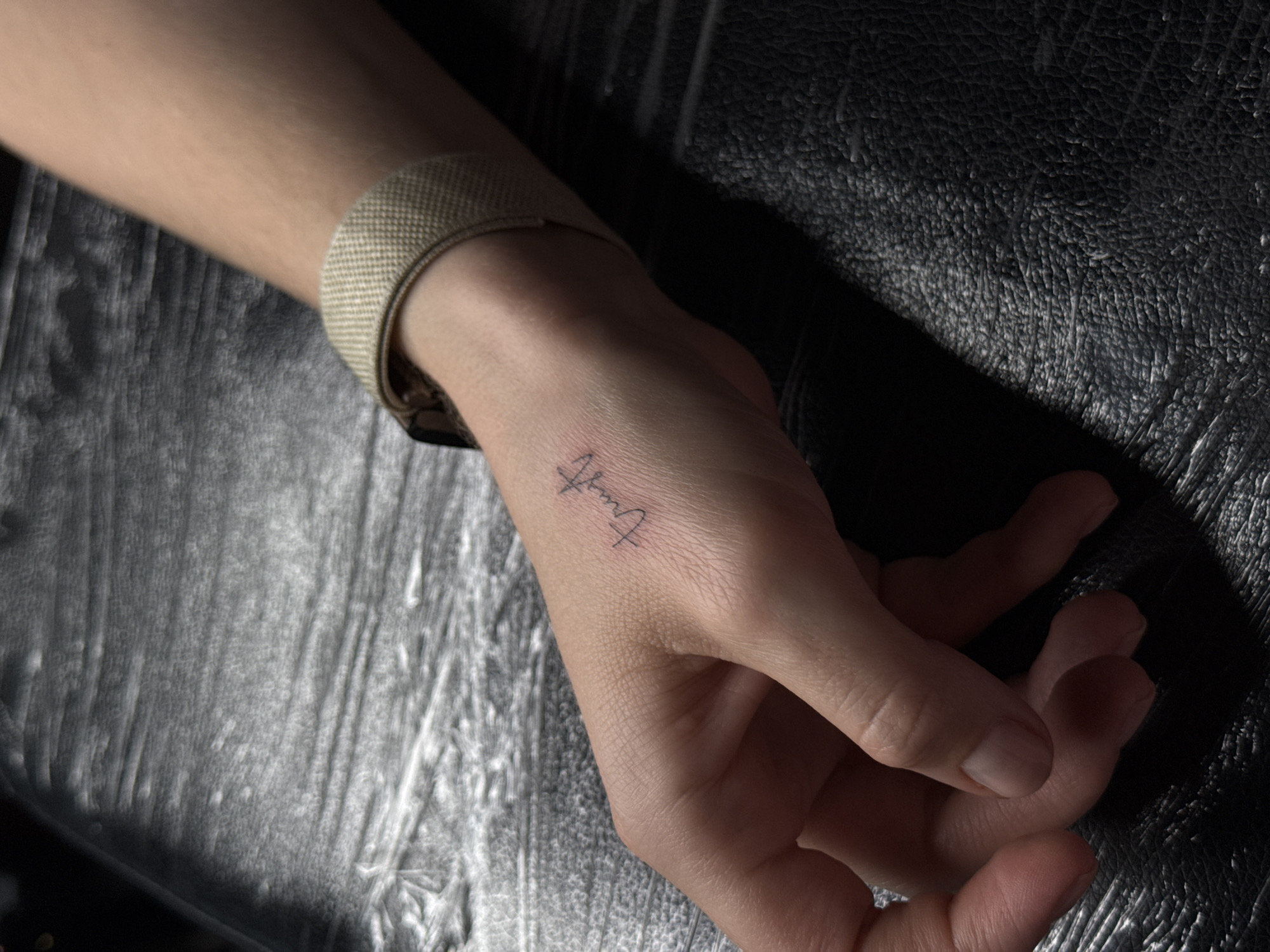A close-up photograph displays a fair-skinned hand with a minimalist tattoo of the word "Truth" in cursive script located on the top of the hand near the wrist. The hand is adorned with a beige watch strap around the wrist. The background consists of two textures: a white, textured material to the left and a dark, grainy surface, potentially leather, to the right. The lighting creates a contrast, emphasizing the texture of the background and casting shadows across the hand. The setting appears to be indoors, possibly a studio or a room with artificial lighting. The location, Khimki, Russia, is not discernible from the image.