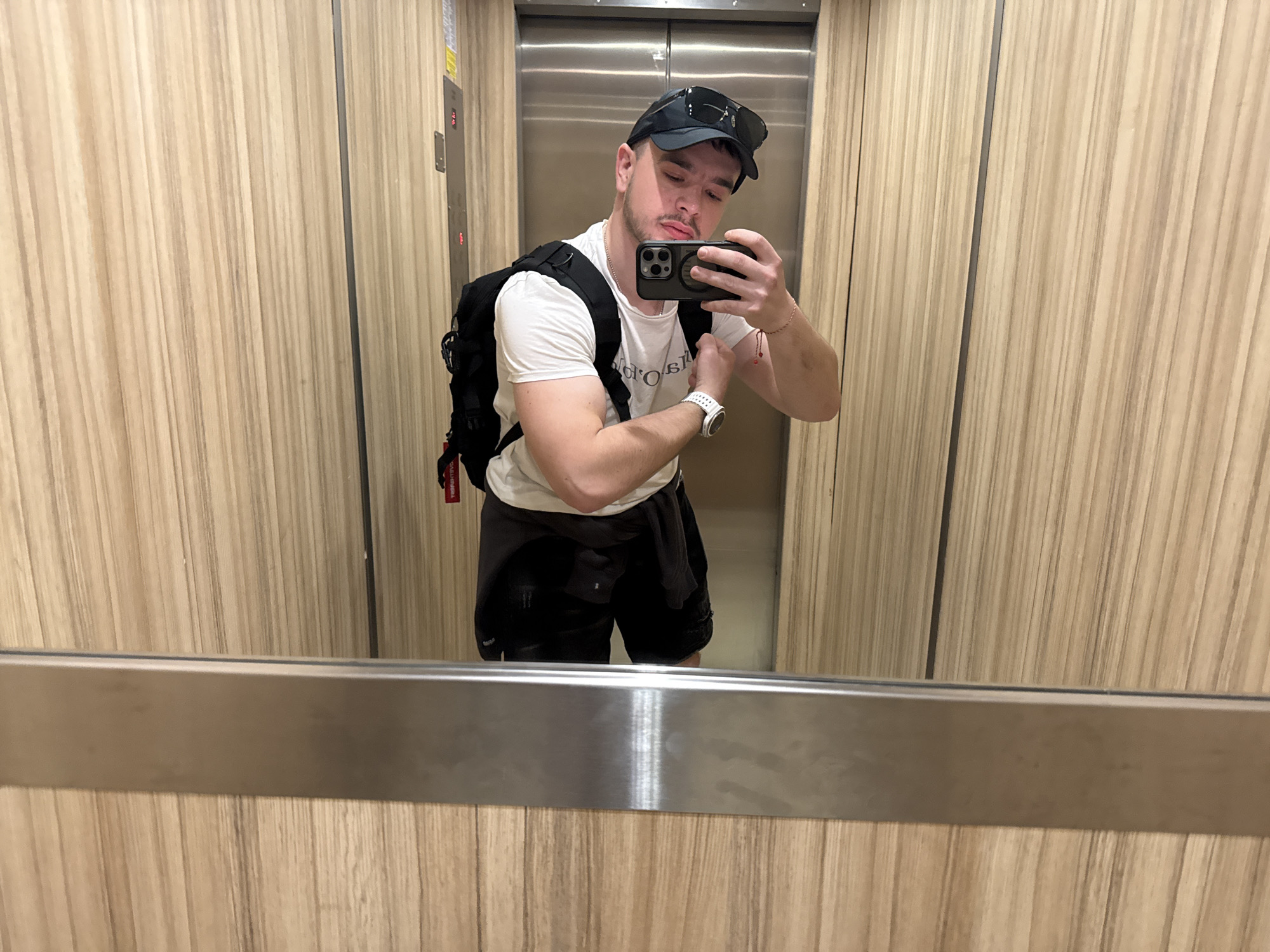 A male with a backpack is taking a selfie in what appears to be an elevator. The male is wearing a white t-shirt, black shorts, a black cap, and a white wristwatch. The elevator's interior is lined with vertically-oriented wood paneling and a stainless steel door. The location is in Sofia, Bulgaria, but there is no specific indicator of this within the image; the determination is based solely on external knowledge.