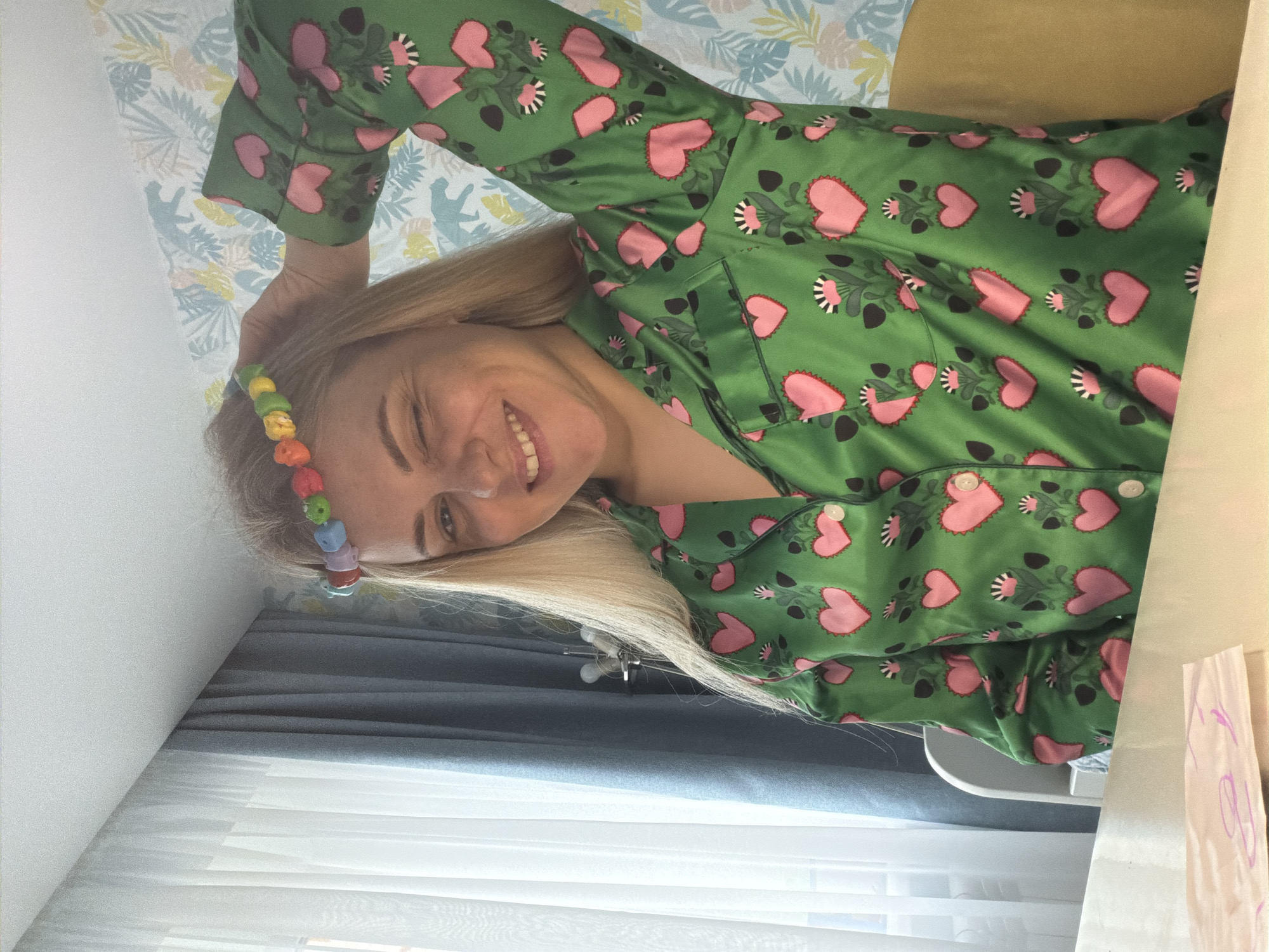 A cheerful blonde woman is captured in a close-up, horizontal shot, smiling brightly and winking with one eye. She is wearing a vibrant green satin pajama top adorned with a pattern of pink hearts and stylized black and white flowers. Her long, blonde hair flows around her, and she has a colorful beaded hair accessory, featuring various pastel hues like yellow, green, blue, purple, orange, and red, in her hair near her temple.
The setting appears to be indoors, likely a bedroom or living room in Votkinsk, Russia. Behind her head is a light-colored wall covered in a whimsical wallpaper featuring subtle leaf and animal silhouettes in muted tones. To her right, a light brown or beige pillow is visible, suggesting she is reclining or resting. In the lower part of the frame, diffused natural light streams through white curtains or blinds, indicating it is daytime. A dark surface, possibly a bed frame or sofa, is behind the pillow. On a surface to the far right, a piece of white paper with some faint, illegible purple writing or drawing can be seen. The woman's expression conveys a happy, relaxed, and playful mood.