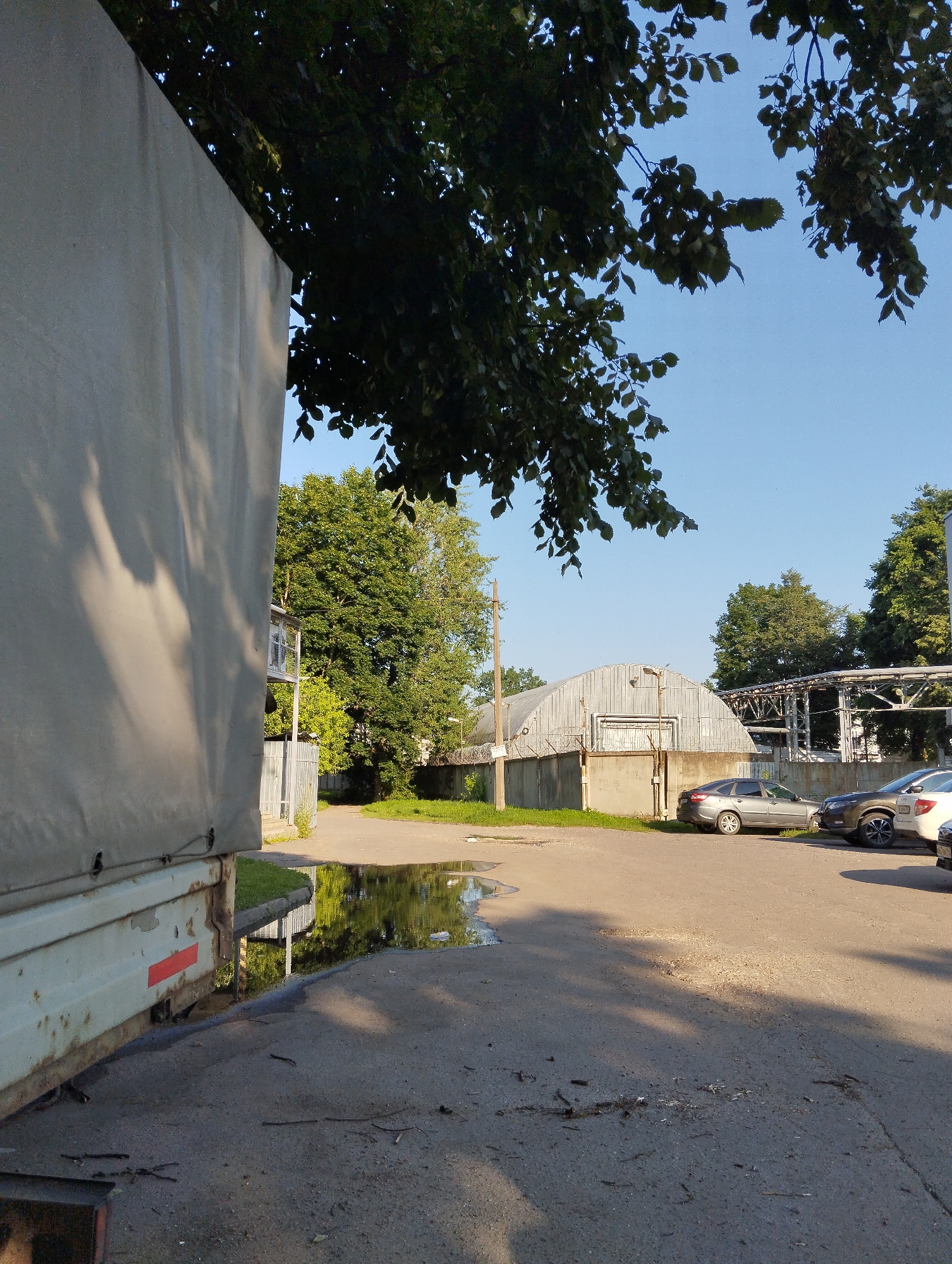 An outdoor, low-angle, eye-level shot captures a parking lot in Kingisepp, Russia, on a clear day. On the left, the side of a large, light-colored truck is visible, showing its corrugated metal construction and some rust. A red reflector is attached to its rear corner. A puddle of water reflects the surrounding environment on the asphalt in the foreground.
In the background, a gray, arched industrial building stands, with a chain-link fence in front of it. Several cars are parked in front of the building, including a dark gray sedan and a dark colored SUV. Trees and a clear blue sky are visible behind the building and vehicles. The overall scene suggests an industrial or commercial area.
