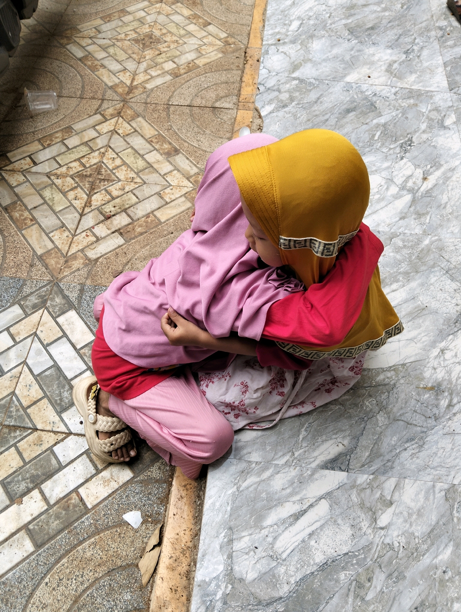Two young children, one wearing a pink hijab and the other a yellow hijab, are embracing each other outdoors. The child in the pink hijab is wearing a pink top and pink pants, and a pair of beige sandals. The child in the yellow hijab is wearing a red long-sleeved shirt and a skirt with a floral pattern. They are positioned on a surface that transitions from patterned tiles to a marble-like floor. The tiled area appears to be a pathway or patio. In the background, a vehicle's tire is partially visible. The scene is lit by natural daylight. The location is identified as Cileungsi, Indonesia.