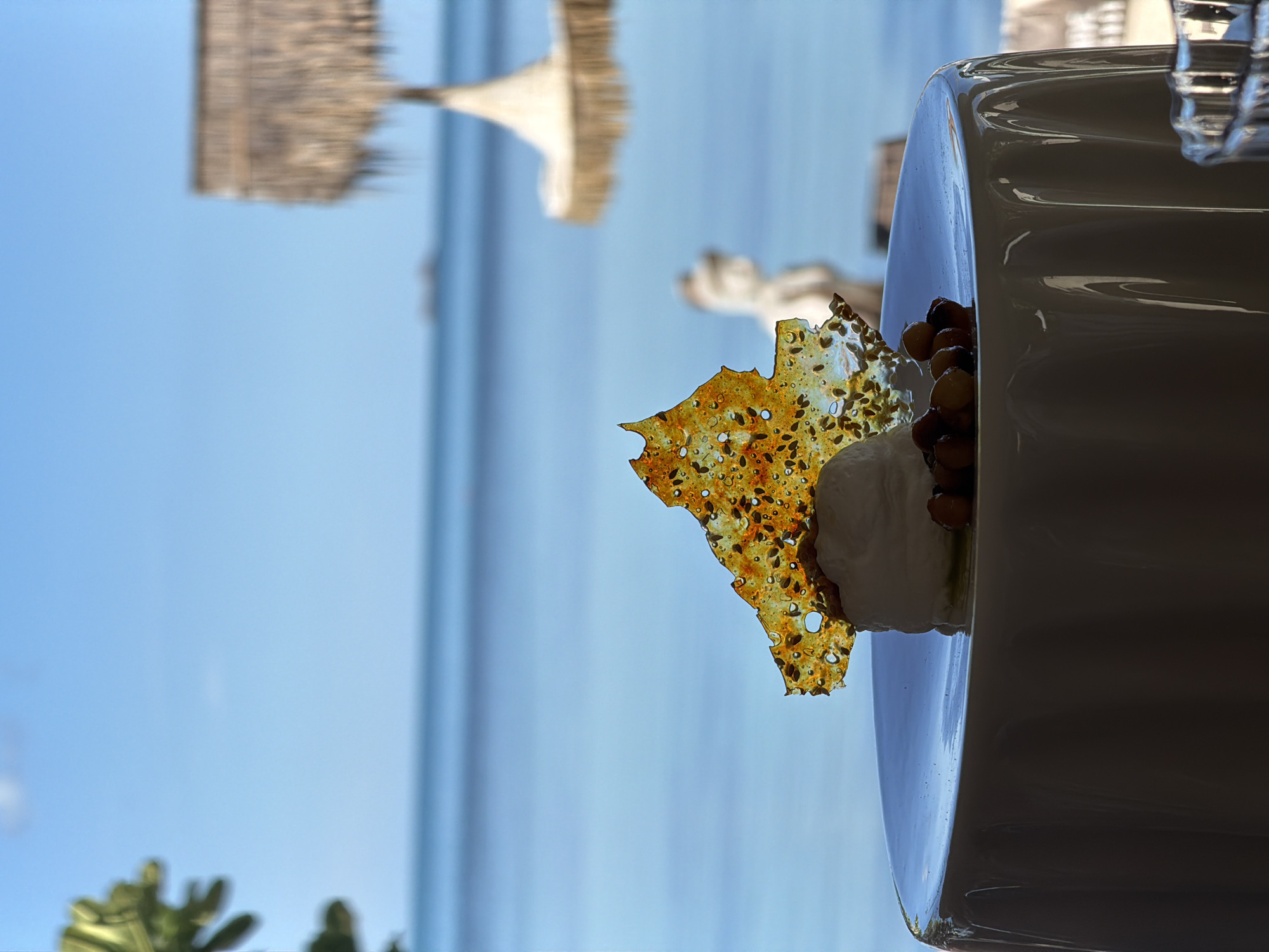 This image, taken in Baan Thong Krut, Thailand, presents a close-up of a dessert served on a white plate, with a backdrop of a bright blue sky and calm water. The dessert appears to be a scoop of white ice cream topped with a brittle, amber-colored confection dotted with seeds, and a cluster of nuts. The scene is likely outdoors, given the clear sky and the suggestion of beach umbrellas or structures in the blurred background. The lighting suggests it is daytime, possibly late morning or early afternoon. The focus is sharp on the dessert, creating a shallow depth of field that blurs the distant scenery, highlighting the culinary subject. The orientation of the image is vertical, with the dessert on the right side of the frame. The overall impression is one of relaxed dining in a tropical setting.