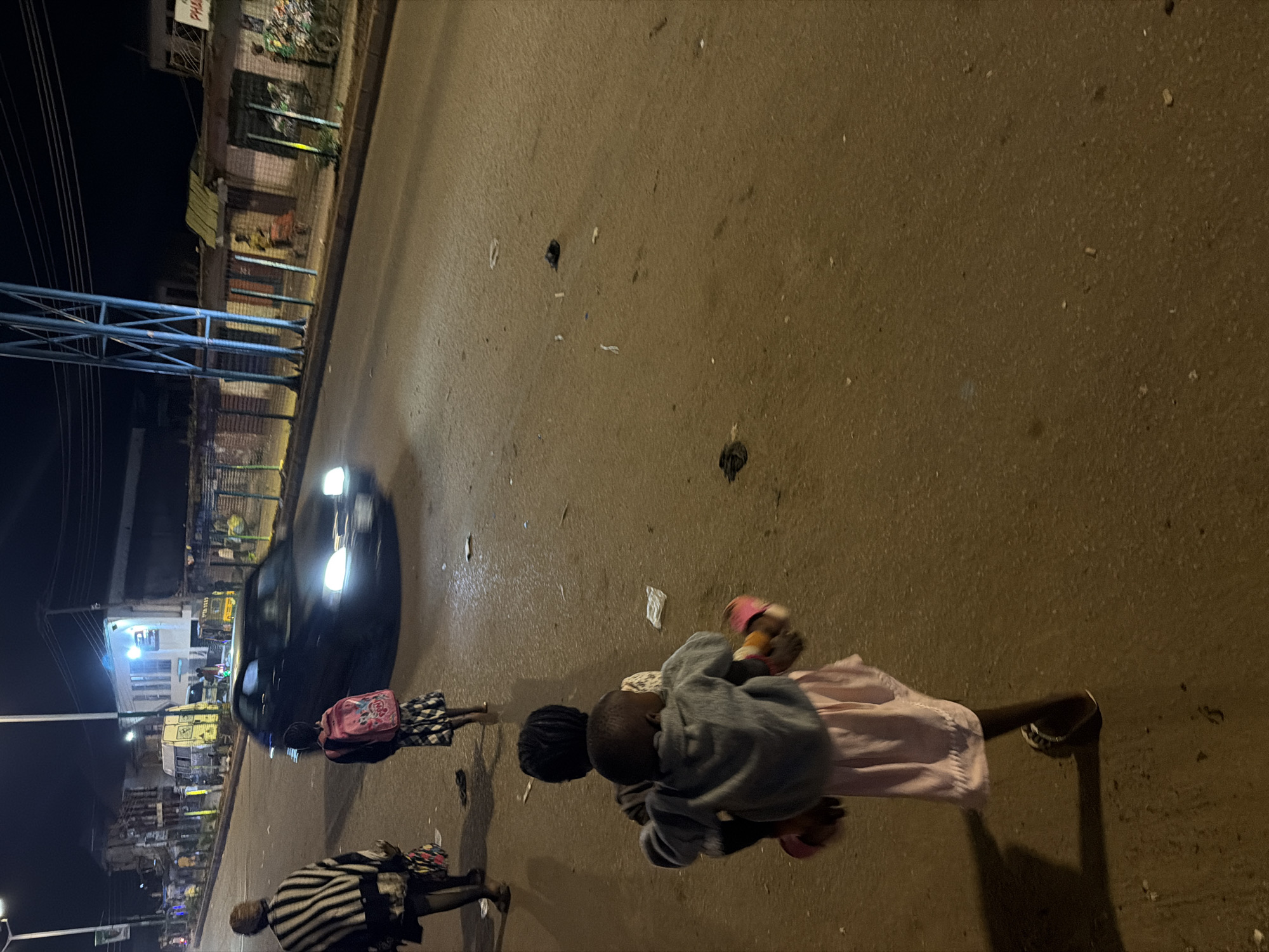 The image depicts a street scene at night in Bukuru, Nigeria. In the foreground, two children are walking away from the camera. The child in front is carrying a younger child on their back, who is wearing a light-colored jacket and a pink skirt. The child in front is wearing a black and white checkered dress. Behind them, another child with a pink backpack and a patterned skirt is walking. Further down the street, an adult wearing a striped shawl is walking. A car with its headlights on is driving past on the right side of the road. The street is paved and appears wet, reflecting the streetlights. The background shows buildings with lights on and some shops with barred windows. Debris, such as plastic bags and wrappers, is scattered on the road. A blue metal structure is visible on the right side of the frame, likely part of an overhead power line or a traffic light pole.