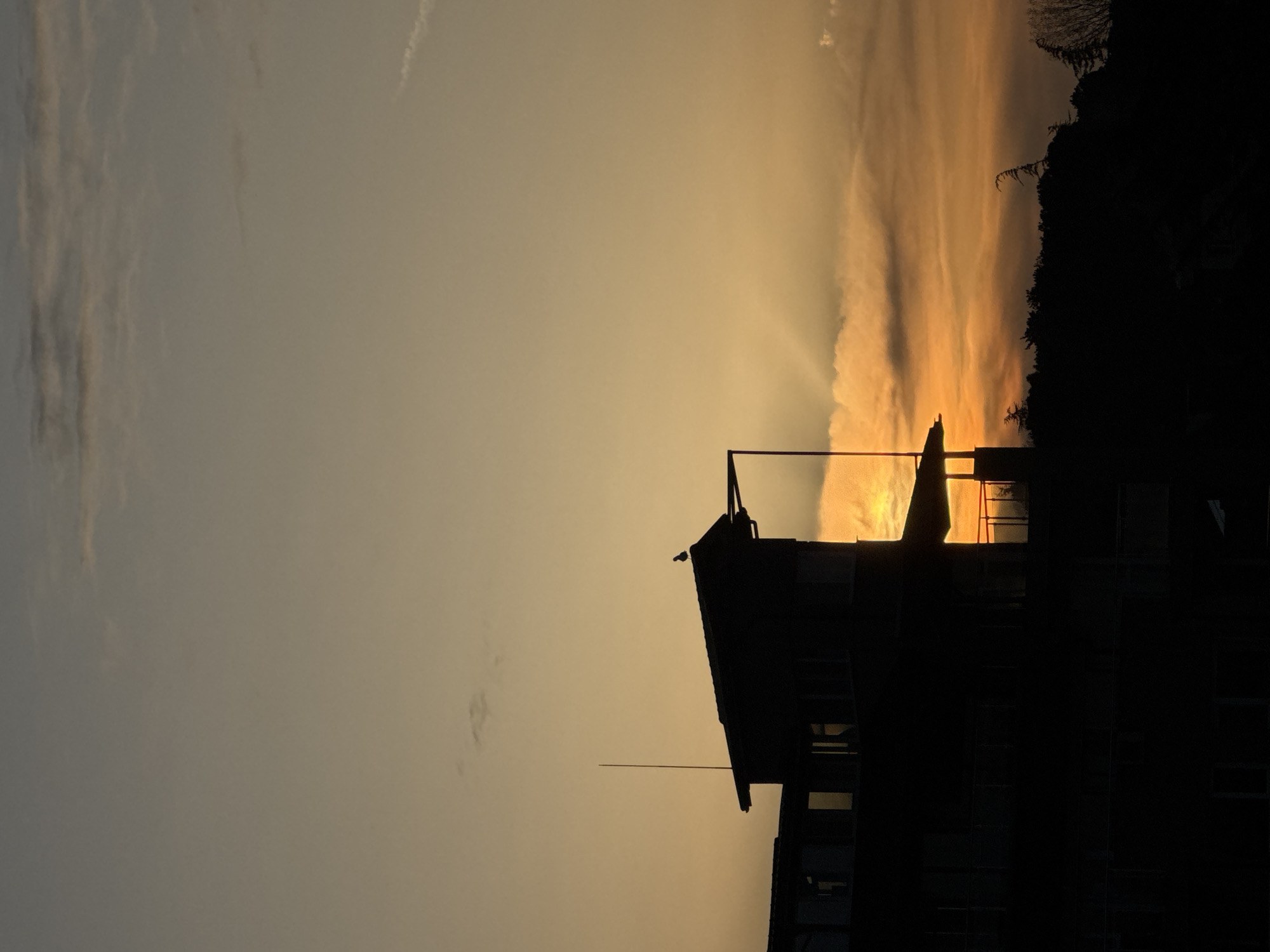 A low-angle, rotated view of a building silhouette against a sunset sky, taken at dusk in Tsarevo, Bulgaria. The sky is a gradient from yellow-orange near the horizon to gray above. A dark treeline is visible on the upper right. The building appears multi-storied, with various structural elements including a roofline, railings, and a tall, thin antenna extending upwards. There is a pole extending from the building's roof, appearing almost to touch the sun. A bird is perched on the roof. The building's features are largely obscured by the dark silhouette.