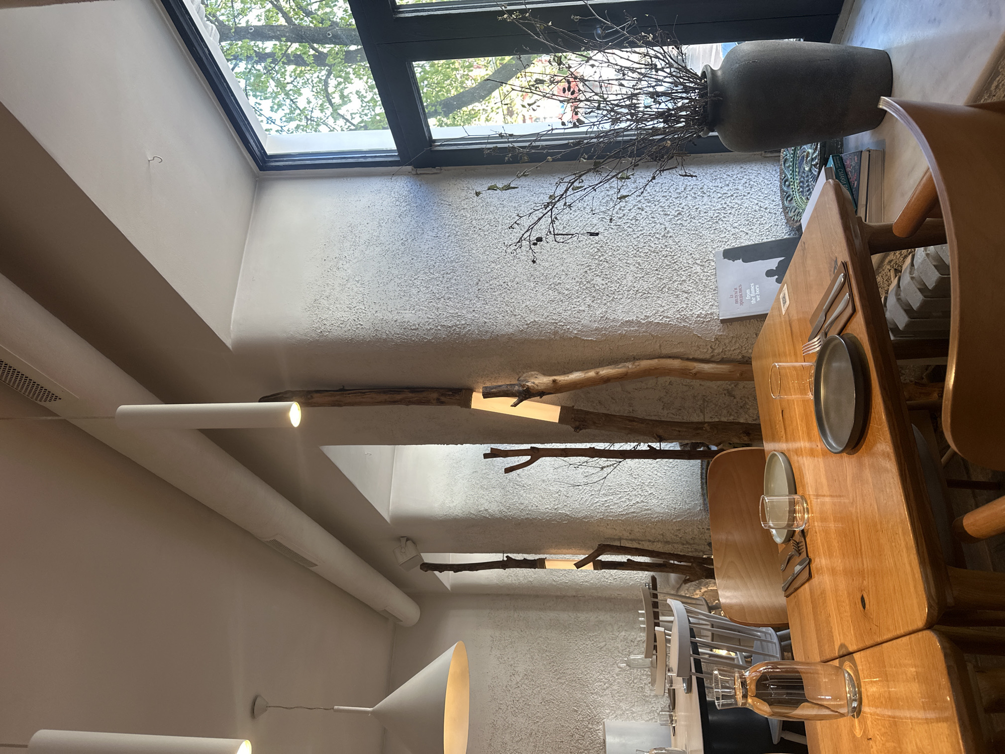 This indoor scene depicts a dining area within a building, potentially a restaurant or cafe, likely located in Kyiv, Ukraine, based on architectural and interior design clues. The space features a high ceiling with a large window letting in natural light. The walls have a textured, possibly stucco-like finish.
A wooden table is the focal point, set with plates, cutlery, and glasses. Chairs, some with wooden frames and others with clear plastic backs, surround the table. A large vase containing dried branches sits near the window.
The ceiling is adorned with linear light fixtures. A decorative element consisting of tree branches is affixed to the wall, possibly integrated with lighting. No people are visible in the scene.