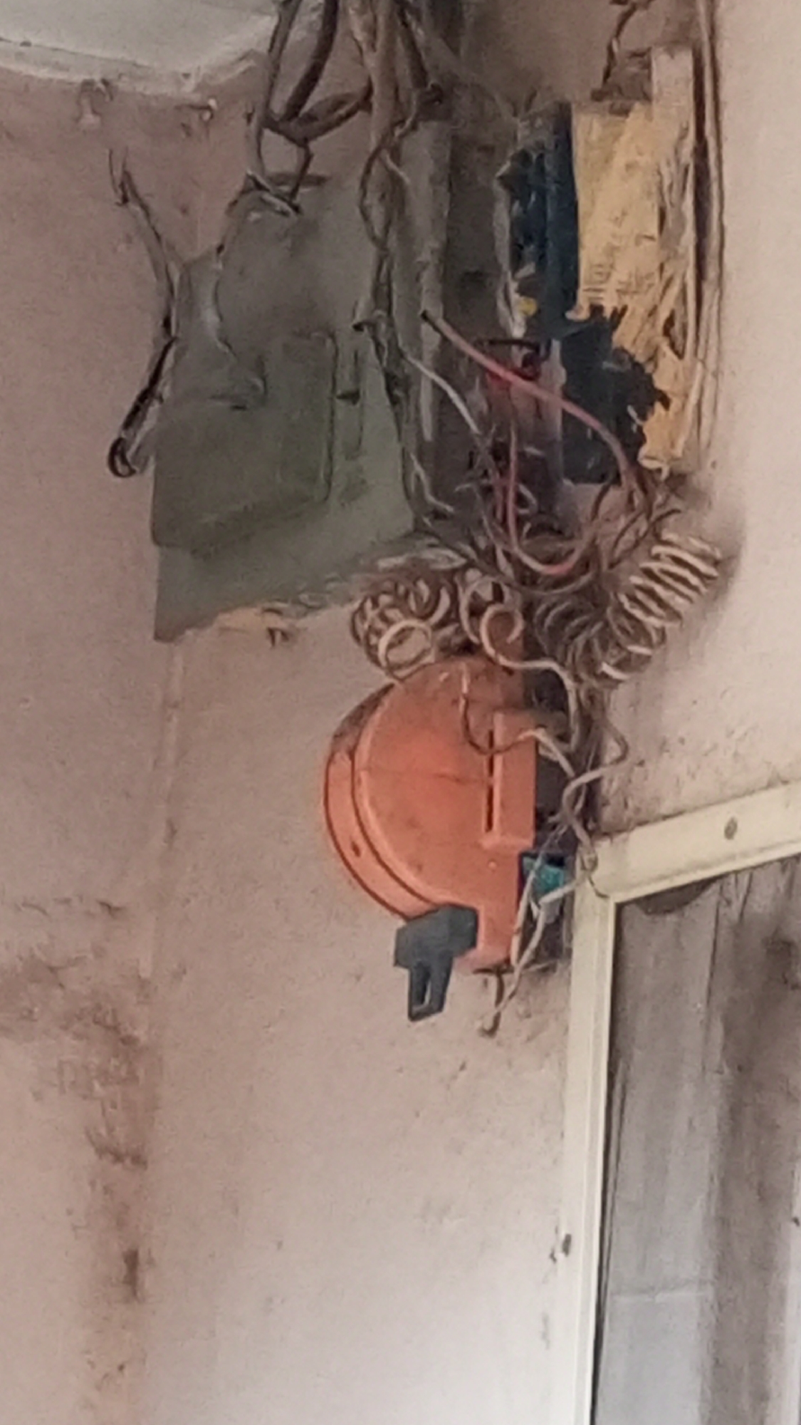 A wall-mounted electrical box and components are visible in a distressed state. The box appears to be exposed with frayed wires and components. An orange circular component with a black switch or handle is attached below the box. The wall is painted a light color with signs of wear and dirt. Part of a window frame with a translucent curtain is visible. Location: Sabon Tasha, Nigeria.