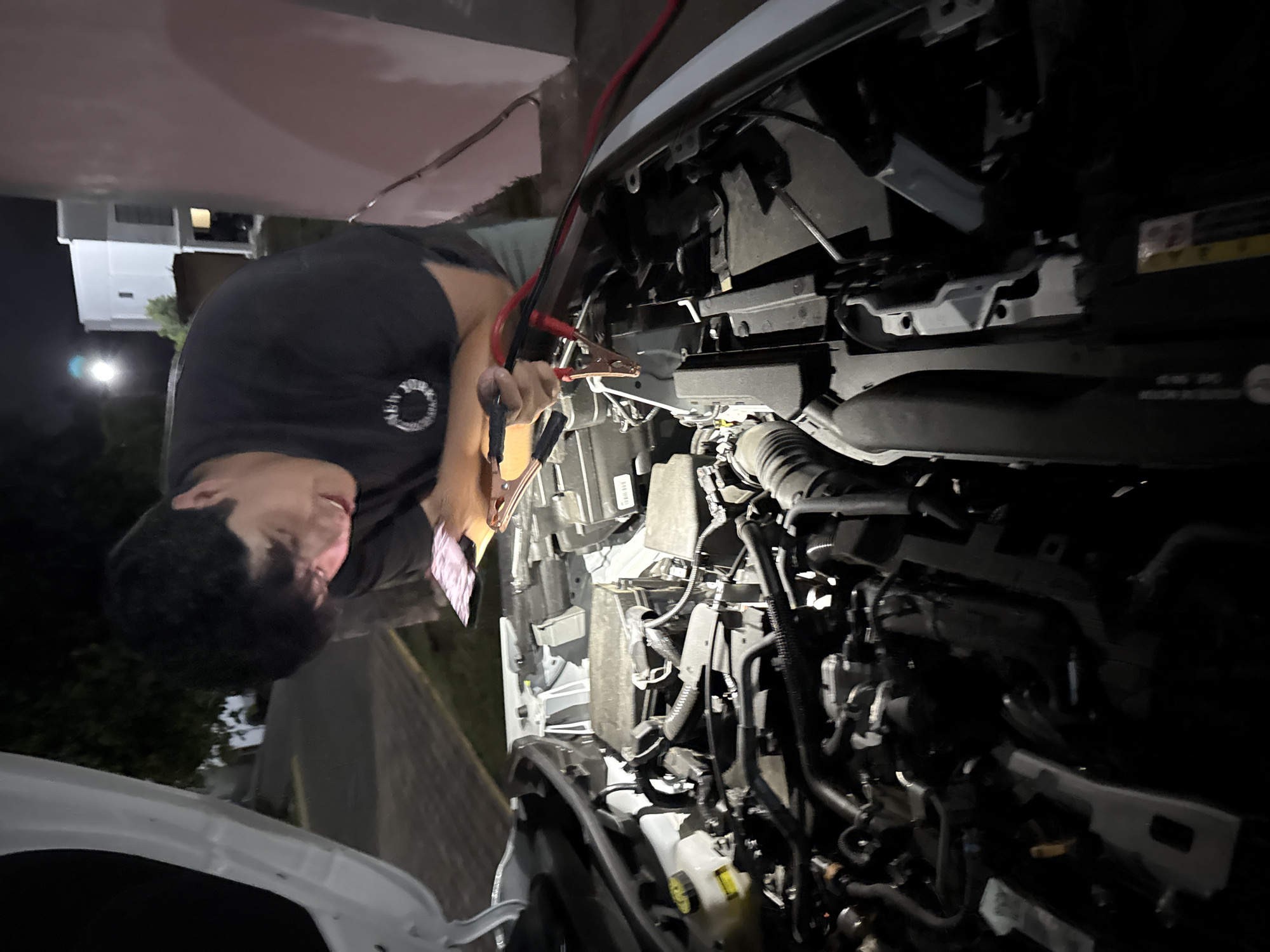 A young man is depicted jump-starting a car at night in Puebla, Mexico. The image is taken from a slightly elevated angle, looking down into the open hood of a white vehicle where the engine bay is exposed.
The central figure, a man with dark hair, is wearing a dark t-shirt with a circular logo on the chest. He is leaning over the front of the car, intently focused on connecting or adjusting jumper cables. One end of the red jumper cable is already attached to a terminal or jump point within the engine bay, and the man is holding the other ends of both the red and black cables in his hands. His expression suggests concentration on the task.
The setting is outdoors at nighttime, with the scene illuminated by an artificial light source, possibly a flashlight or nearby streetlights, casting some shadows. In the background, a residential structure with illuminated windows and dark trees are visible against the night sky, indicating a suburban or urban environment. A paved road or driveway is also discernible. The activity suggests a car trouble situation requiring a jump-start. No specific legible text or branding is clearly visible on the car parts or the man's clothing due to resolution and lighting.