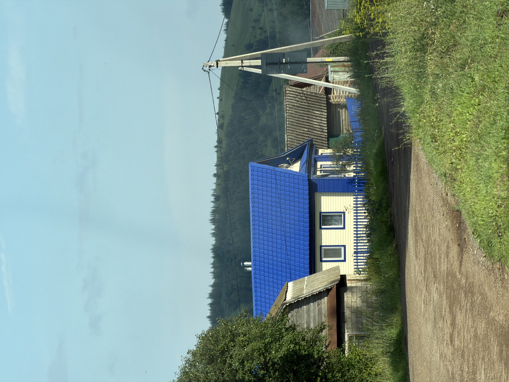 The image shows a rural village scene. In the foreground, a dirt road curves to the right, bordered by green grass and vegetation. Several houses are visible behind the road. One house in the center has a bright blue roof and yellow siding with white trim around two windows. A blue picket fence runs in front of this house. To the left of this house is another structure with a weathered wooden roof and brick foundation. Further to the right, another house with a corrugated roof is visible. A utility pole with wires and what appears to be a sign is situated to the right of the houses. In the background, rolling green hills covered in trees rise against a pale blue sky with faint clouds. The overall impression is that of a quiet, possibly remote, settlement on a clear day.