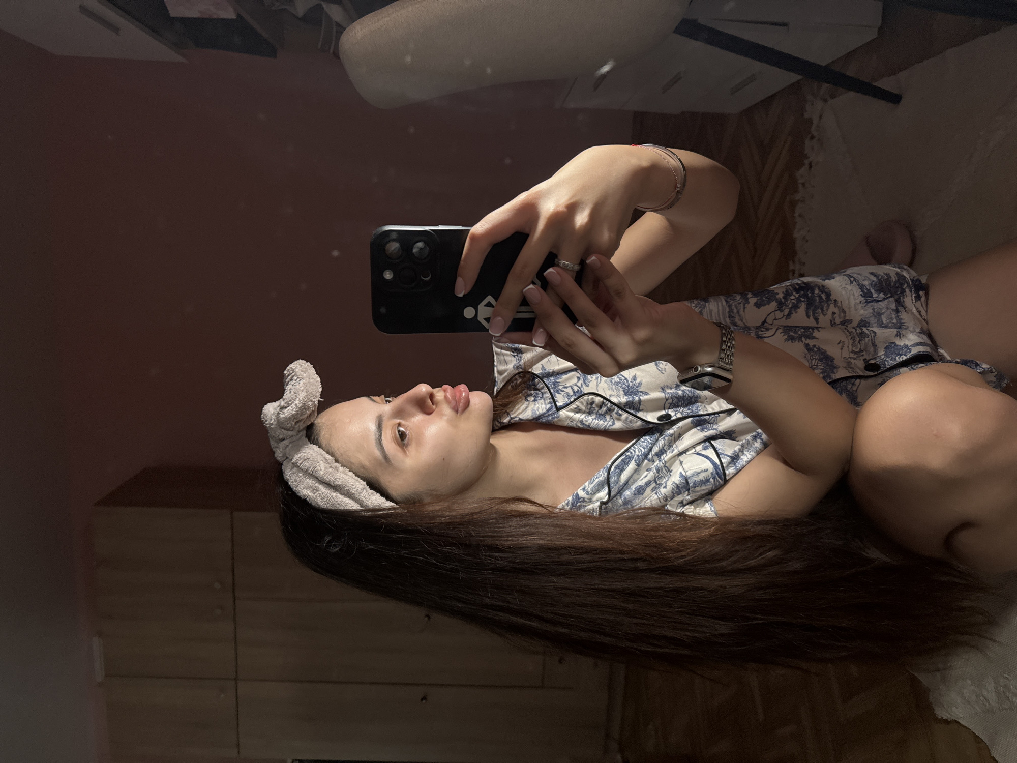 A young woman with long, dark brown hair and a light-colored headband is taking a selfie in a bedroom. She is wearing blue and white patterned pajamas and is kneeling on the floor, holding a smartphone in her right hand. Her left arm is resting on her leg. The room appears to be dimly lit, with wooden cabinets visible in the background. The flooring is a mix of wooden parquet and a light-colored rug. A chair and a dresser are visible in the mirror's reflection. The image was captured indoors, likely in a residential setting. It is not possible to determine the specific location within Plovdiv, Bulgaria from the visual information provided.
