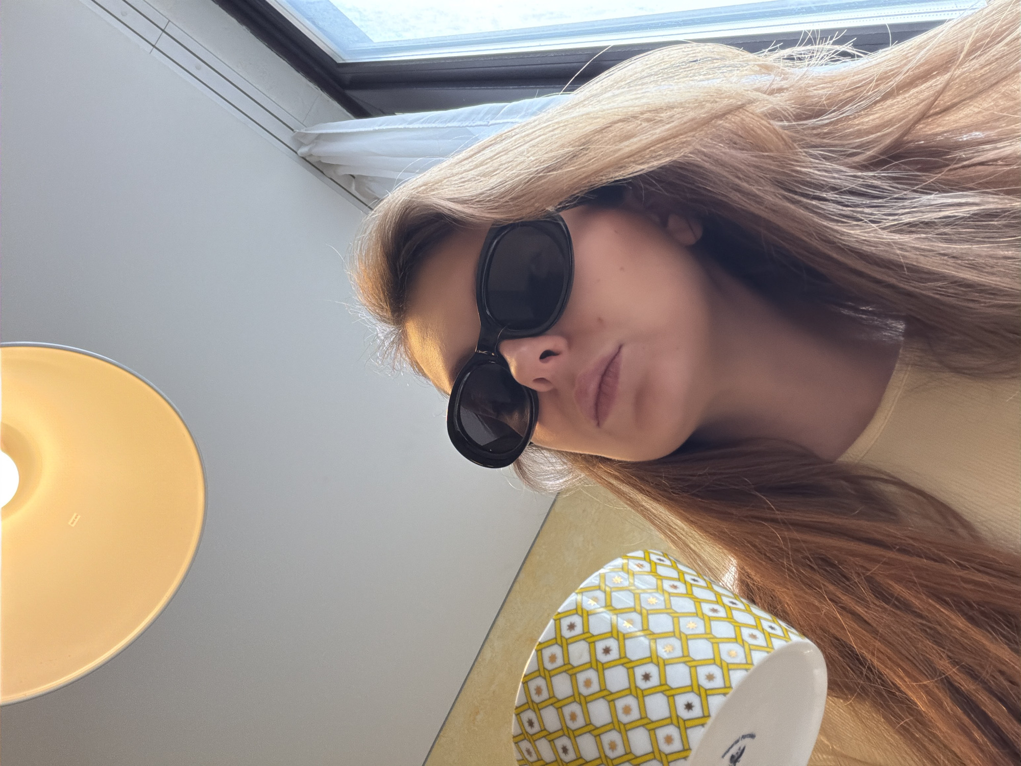 A woman with long, light brown hair and wearing dark sunglasses looks down into the camera. She is holding a patterned cup, predominantly yellow and white, with gold accents. A bright yellow lamp is visible to the left. A window is in the top left corner. The location is not determinable from the image.