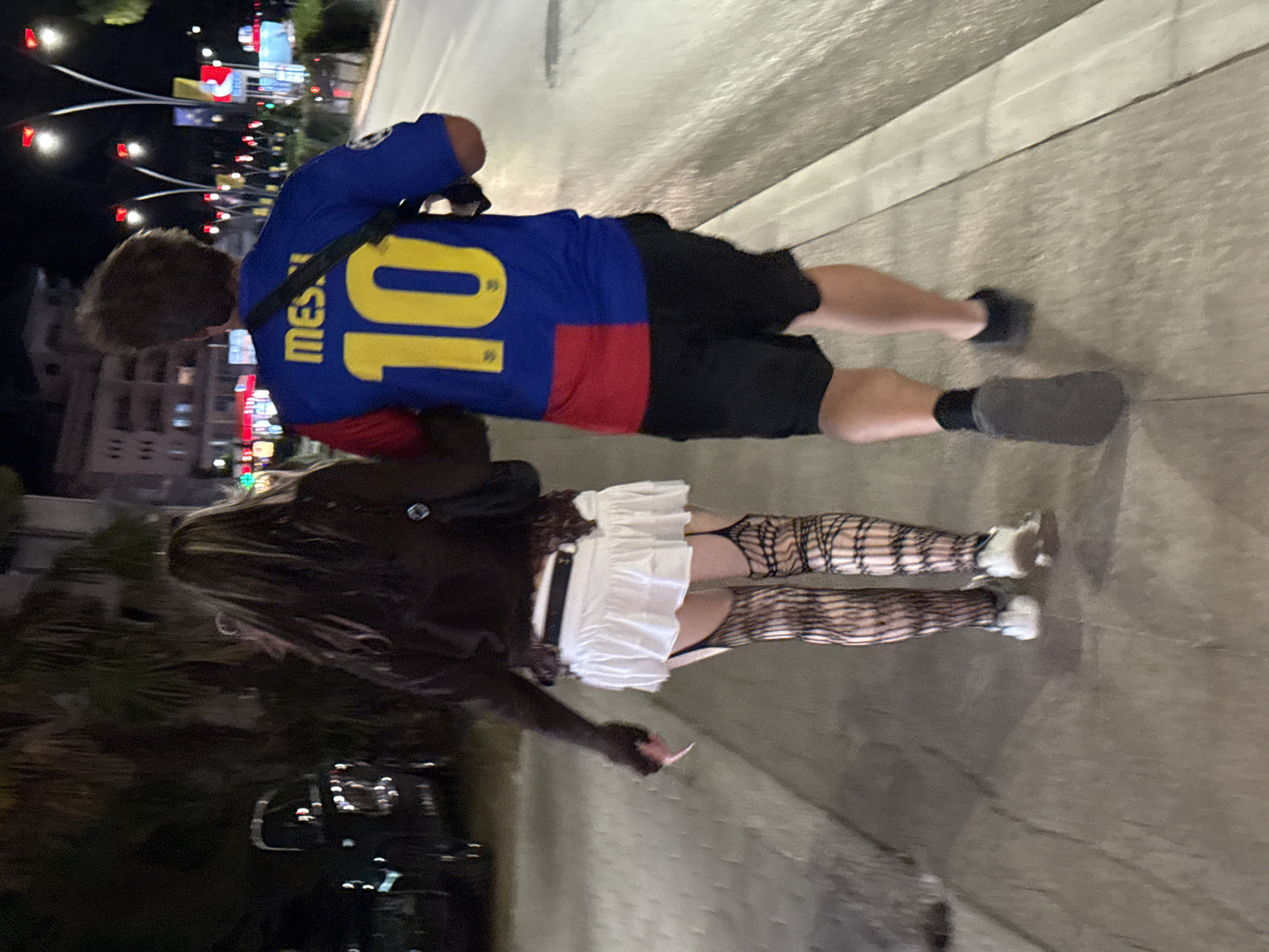 The image shows a couple walking on a sidewalk at night in Budva, Montenegro. The man is wearing a blue and red soccer jersey with "MESSI" and the number "10" on the back, and black shorts. The woman is wearing a white ruffled skirt, a dark brown top, fishnet stockings, and sneakers. They are walking away from the camera, towards a street lined with palm trees and illuminated buildings. The atmosphere suggests a casual evening stroll in a tourist area.
