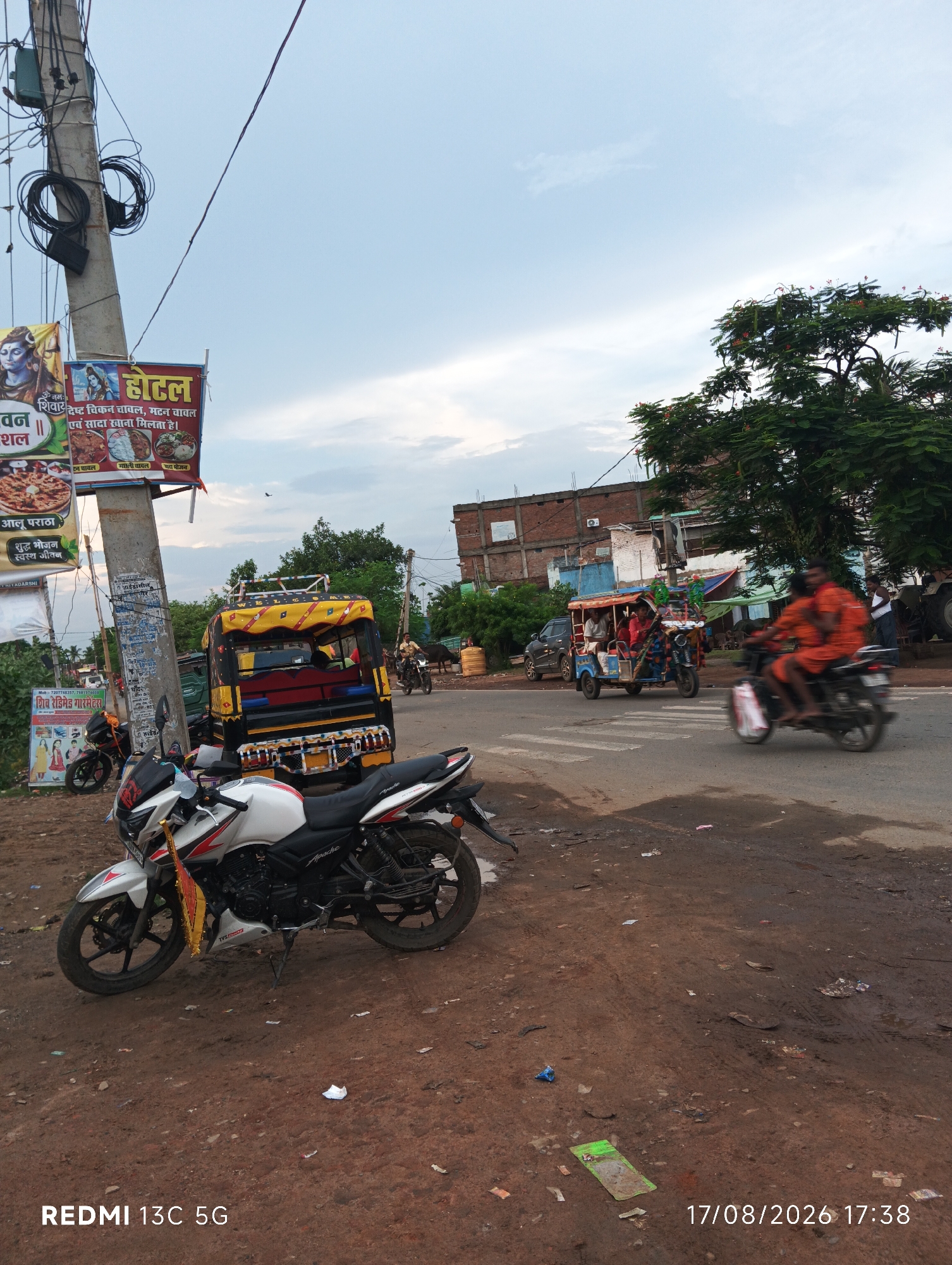 The image shows a street scene in Bariyarpur, India. A white motorcycle is parked on the side of a dirt road, with a pole and wires to its left. Behind the motorcycle, a yellow and black auto-rickshaw is parked. In the background, other vehicles, including another auto-rickshaw with passengers and a motorcycle with two people, are moving on the paved road. Several buildings, including a brick structure, are visible in the distance, along with trees. The sky is cloudy, and the overall lighting suggests it is daytime. The dirt road is littered with debris. Signs in Hindi are visible on the pole and on a banner to the left of the motorcycle. A timestamp and "REDMI 13C 5G" are visible at the bottom of the image.