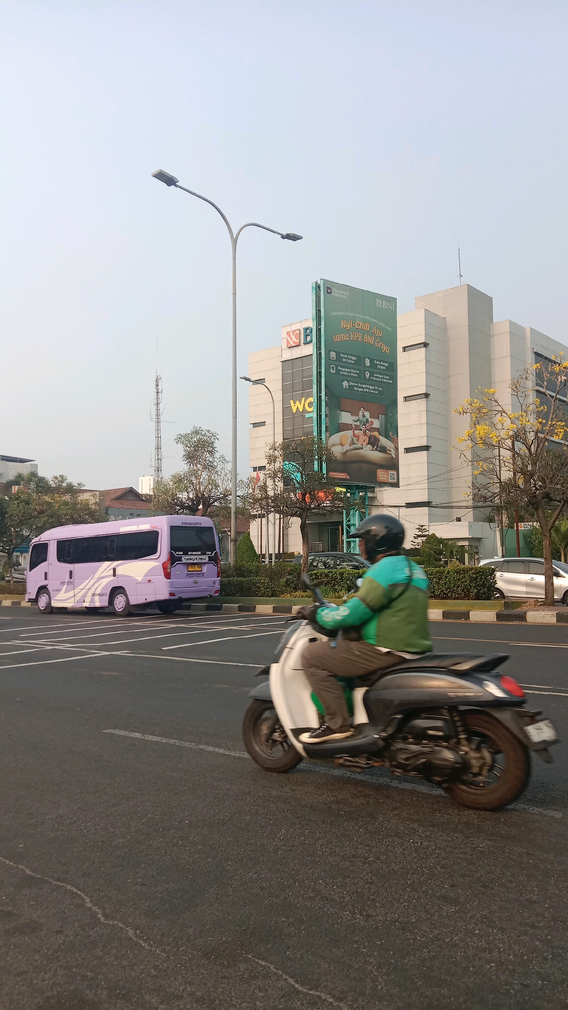 The image depicts a street scene in Bekasi, Indonesia, during daylight. In the foreground, a person wearing a green jacket and a helmet is riding a white and grey scooter. They are moving along a paved road marked with white lines. In the middle ground, a purple minivan with "PARIWISATA" written on its rear is parked on the left side of the road. Further in the background, a modern building with a large billboard displaying an advertisement for BNI Griya is visible. The billboard features text in Indonesian and an image of a family. The sky is clear and pale. The scene includes streetlights and trees along the roadside.