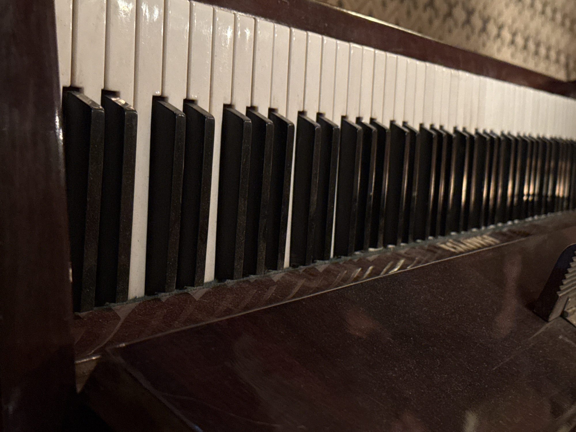 The image shows a close-up of a piano keyboard, revealing the white and black keys in sharp focus. The piano is a dark, glossy wood finish, appearing well-used with some dust visible on its surface. The keys themselves show signs of wear. The perspective is angled, making the keys in the foreground appear larger and gradually diminishing in size towards the background, indicating the depth of the keyboard. The setting is indoors, suggested by the presence of patterned wallpaper visible in the blurry background. There is no indication in the image that identifies the location as Tel-Aviv, Israel.