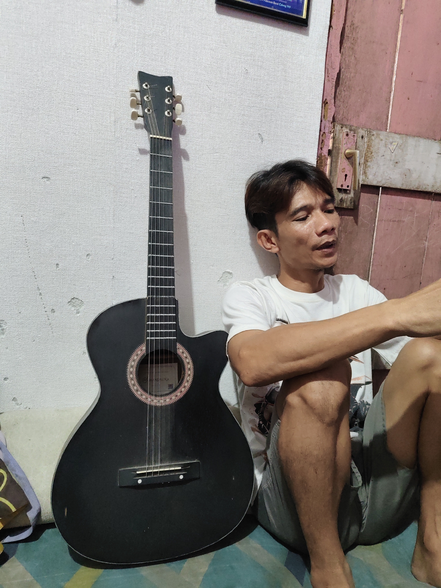 An Indonesian man sits on the floor next to a black acoustic guitar, leaning against a textured white wall. He wears a white t-shirt and grey shorts. The guitar is positioned upright, leaning against the wall to his left. The background features a wooden door with a rustic finish and a faded sign hanging on the wall above the man's head. The floor is covered with a patterned mat. The scene appears to be indoors, possibly in a residential setting within South Jakarta, Indonesia, given the style of the door and wall texture, common in older Indonesian buildings.