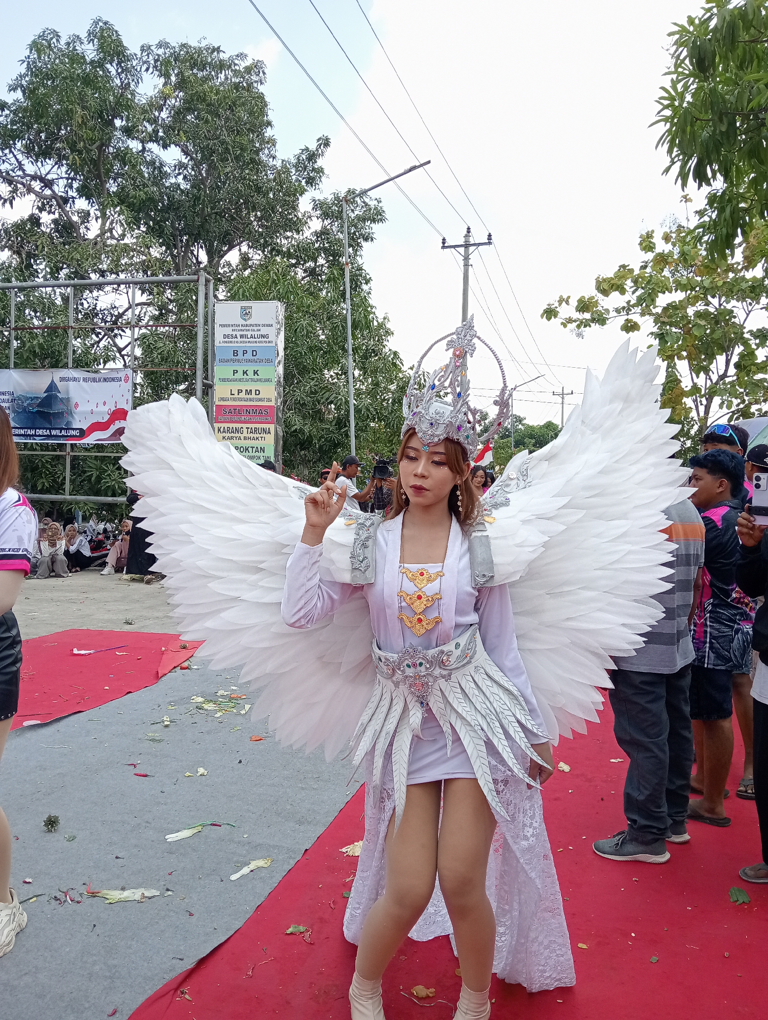 A young woman in elaborate white angel wings and a silver crown stands on a red carpet in Wilalung, Indonesia. She is wearing a white dress with gold embellishments and a flowing white train. Her makeup is dramatic, with bold eyeliner and lip color. She is posing with one hand raised, her index finger pointed upwards. Several people are visible in the background, some watching the woman and others taking photos. A sign in Indonesian, likely an official notice, is posted to the left of the woman. The scene appears to be an outdoor event, possibly a parade or festival. The ground is littered with confetti and debris.