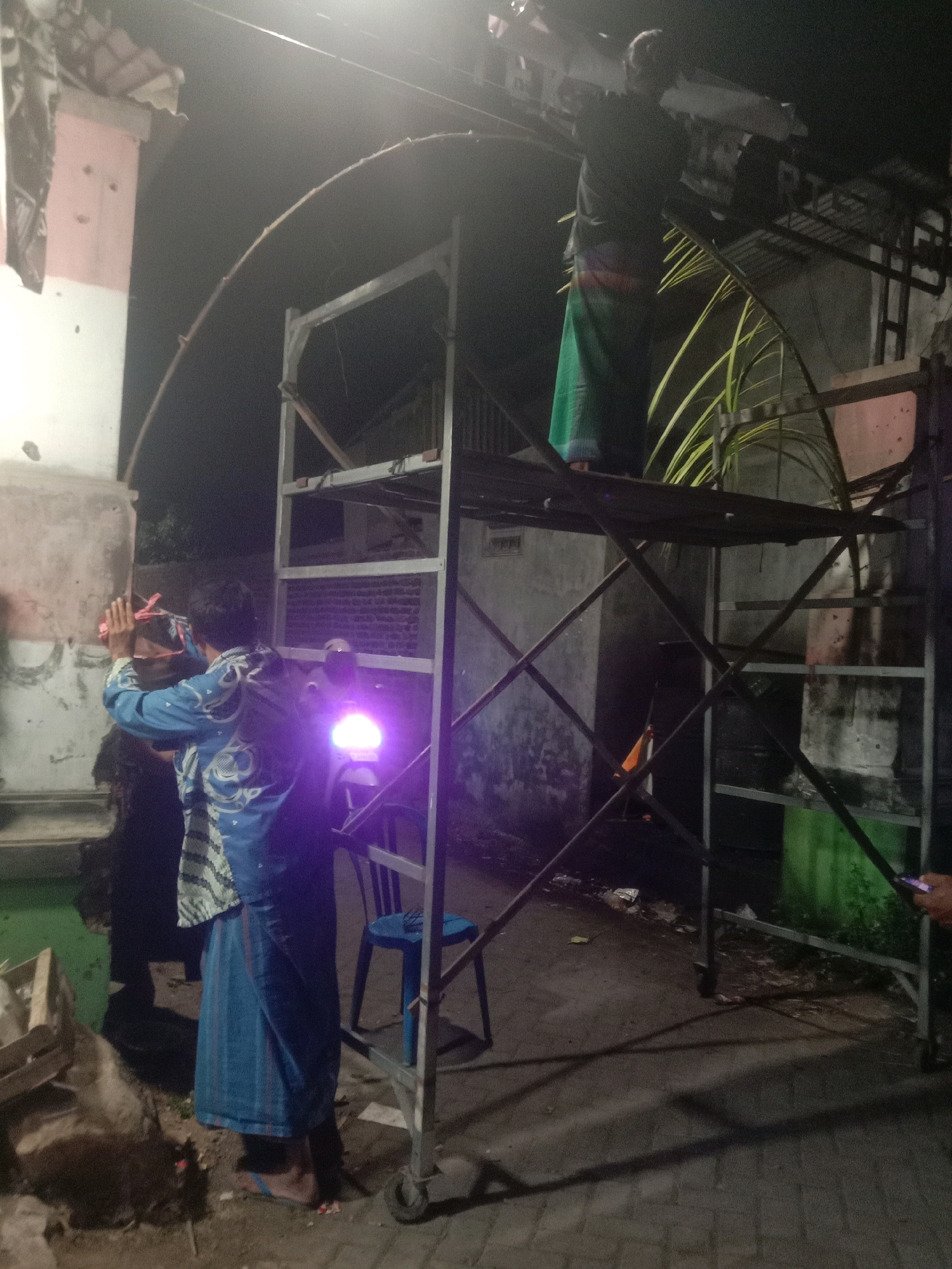 The image depicts a nighttime scene on a paved street in Karanganyar, Indonesia. In the foreground, a person wearing a blue and white patterned shirt and a blue sarong stands near a scaffolding structure. They are holding a bright purple light, possibly a flashlight, which illuminates the area and casts a shadow on the ground. To the left of the scaffolding, a wooden crate is visible.
On top of the scaffolding, another person is positioned, wearing a dark shirt and a green and white sarong. This individual appears to be working on a long, curved object, possibly a decoration or structure, which extends from the scaffolding towards the left side of the image. The scaffolding itself is made of metal and has multiple platforms and diagonal supports.
In the background, a building with a pink and white facade is visible, with some circular indentations on its surface. The overall environment suggests an outdoor setting, possibly a public area or a residential street where preparations for an event or construction are taking place. The lighting is dim, with the purple light from the flashlight and some ambient illumination from the surroundings.